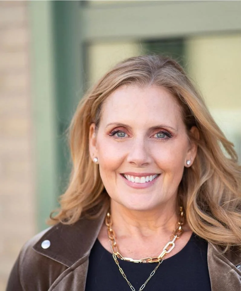 A woman with blonde hair and blue eyes smiling, wearing pearl earrings, a black top, a brown jacket, and gold jewelry, outside in front of a window.