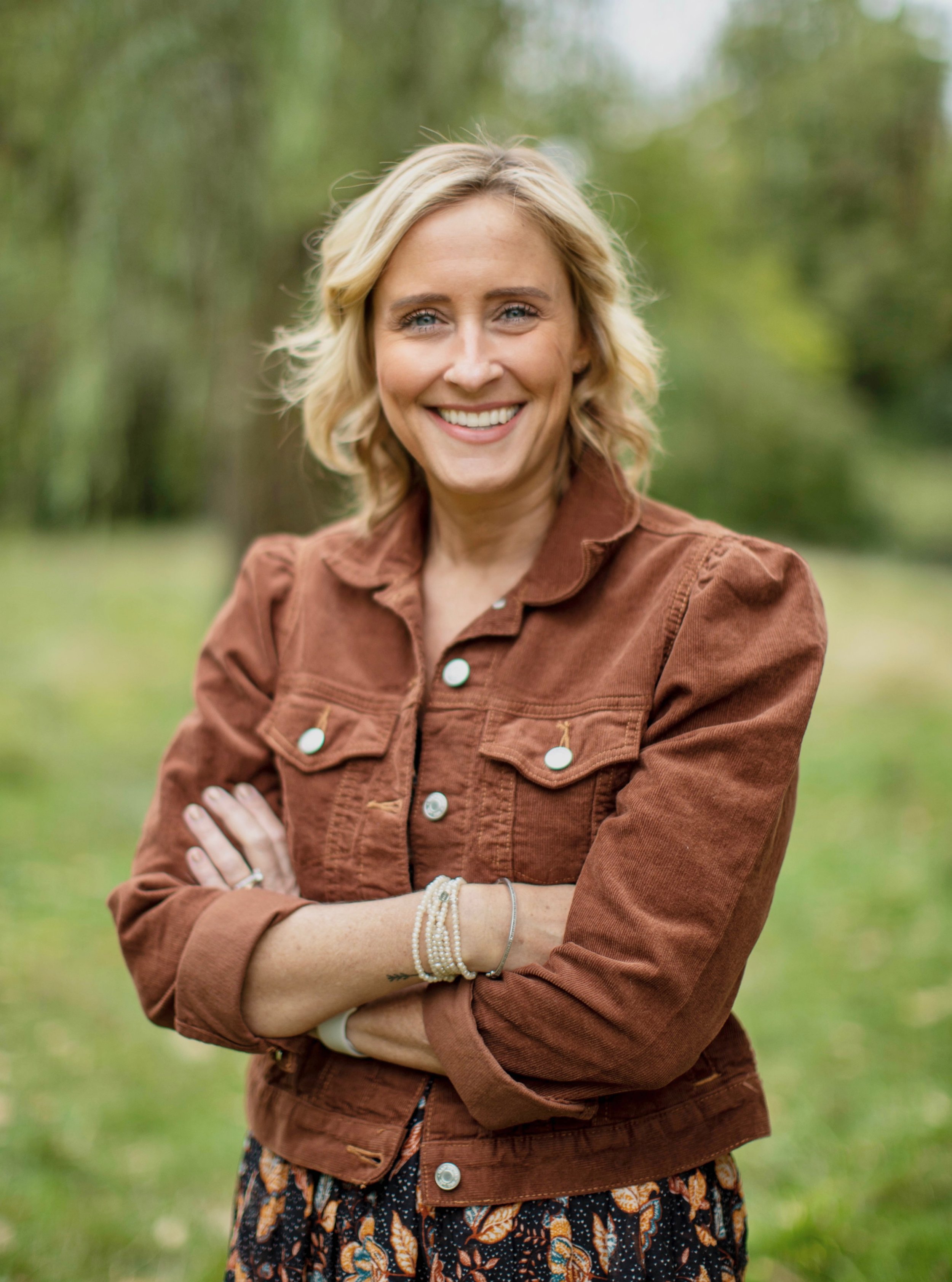 A smiling woman with blonde hair in a brown jacket standing outdoors in a park with green trees in the background.