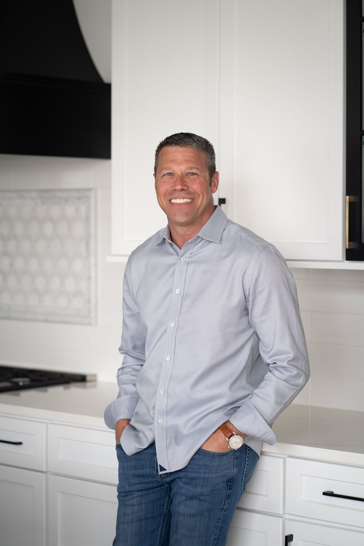 A smiling man in a light gray button-up shirt and jeans standing in a modern white kitchen with hands in pockets.