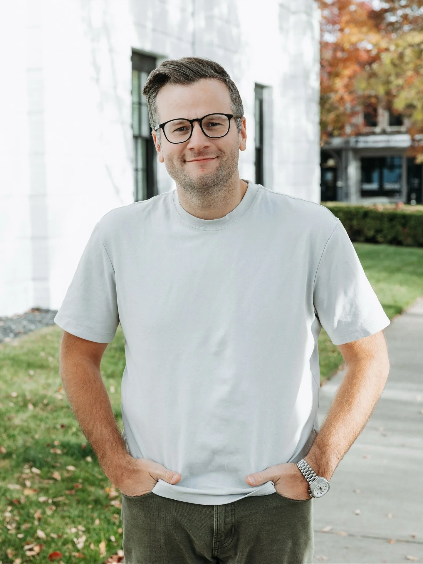 A man with glasses wearing a light gray T-shirt and a watch on his left wrist, standing outdoors with hands in his pockets, smiling at the camera.