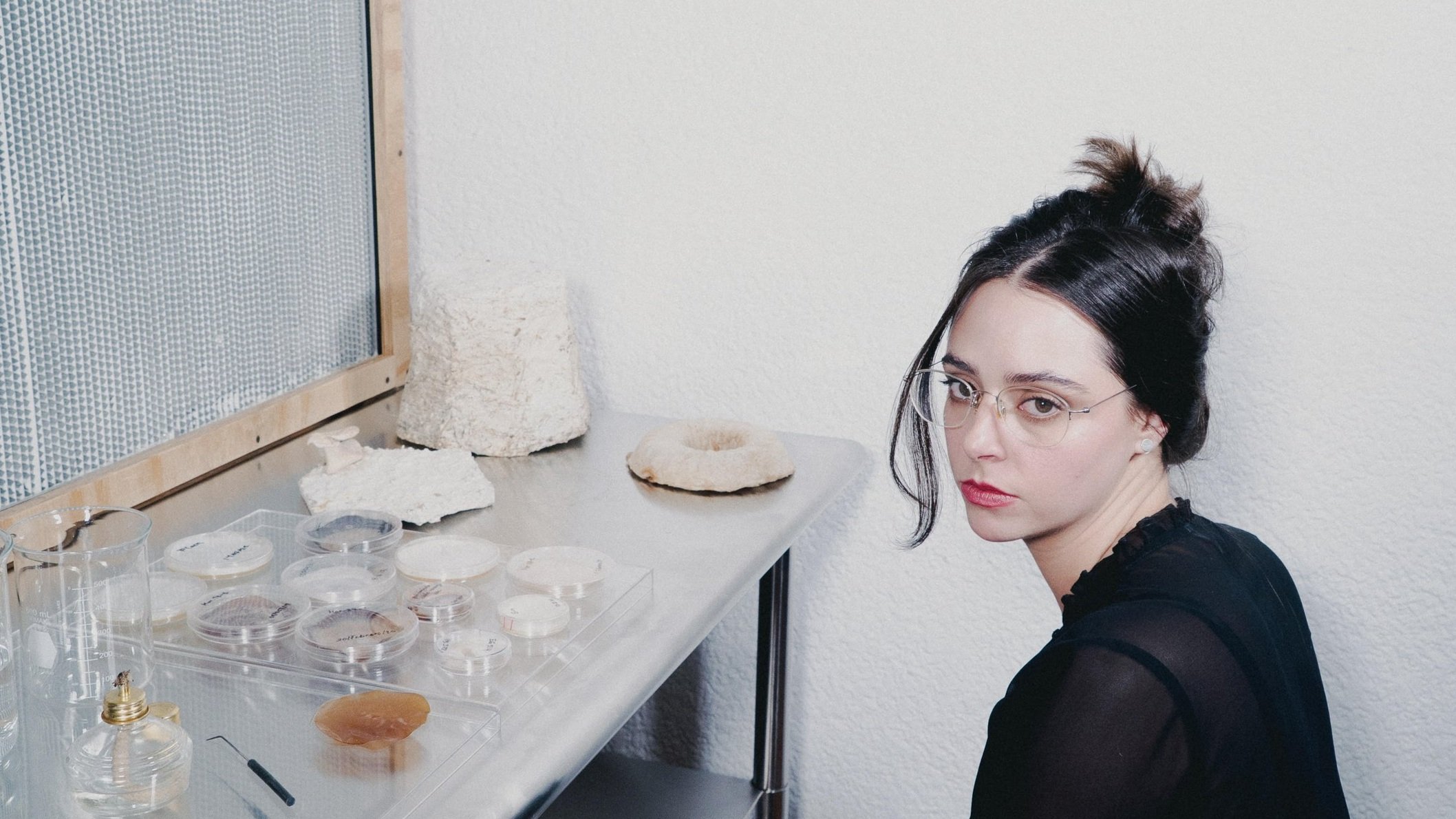 María Monteys, co-founder of Monterr, with dark hair, glasses, and light skin, seated at a laboratory table surrounded by scientific equipment including beakers, petri dishes, and mushroom samples.