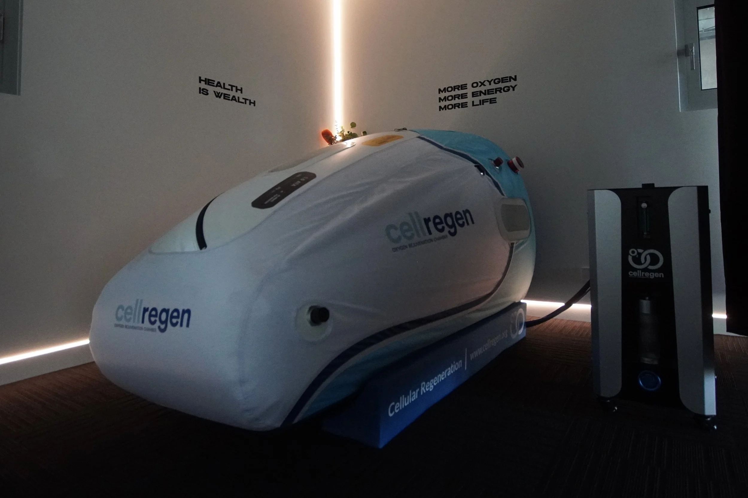A portable hyperbaric oxygen therapy chamber labeled CellRegen, designed for cellular regeneration therapy, resting on the floor in a room with gray tiled flooring, a wall-mounted TV, and informational posters.