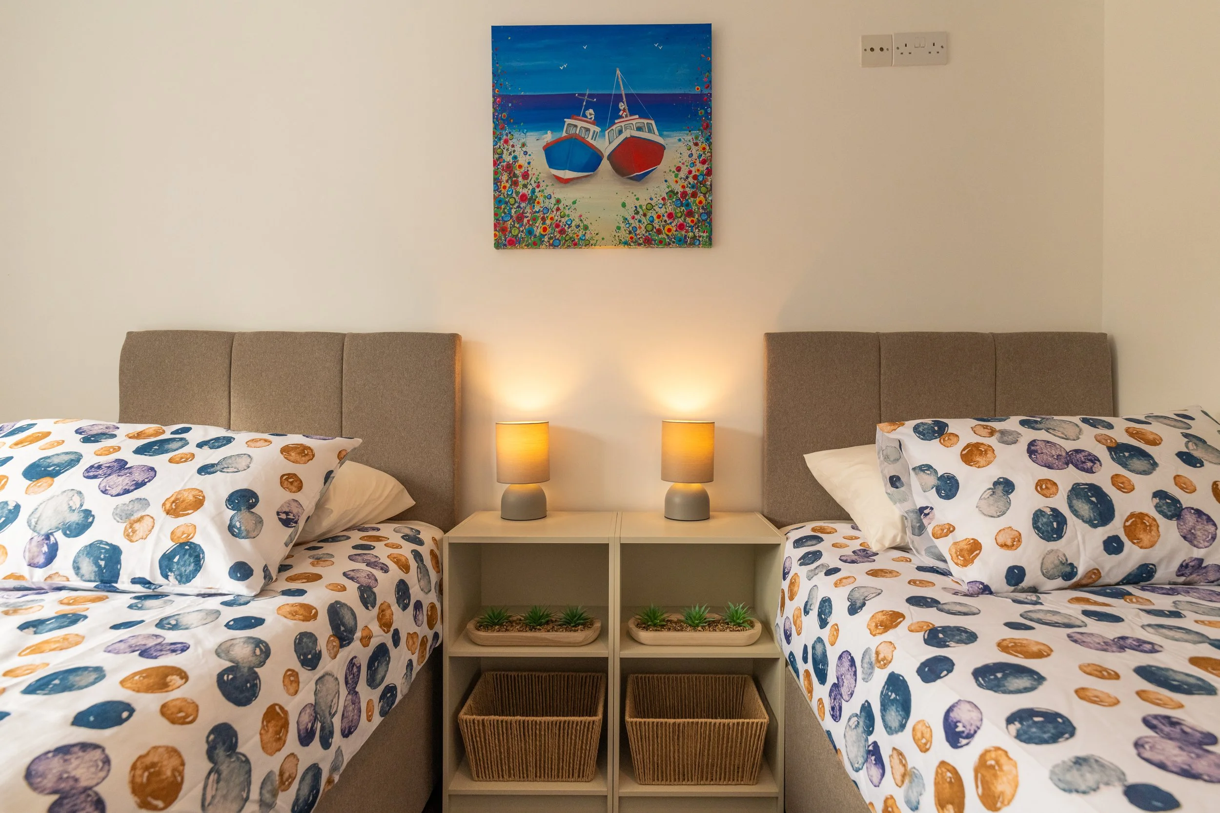 Twin beds with colorful polka dot bedding in a room with a shared nightstand, two lamps, and a beach-themed painting of boats hanging on the wall.