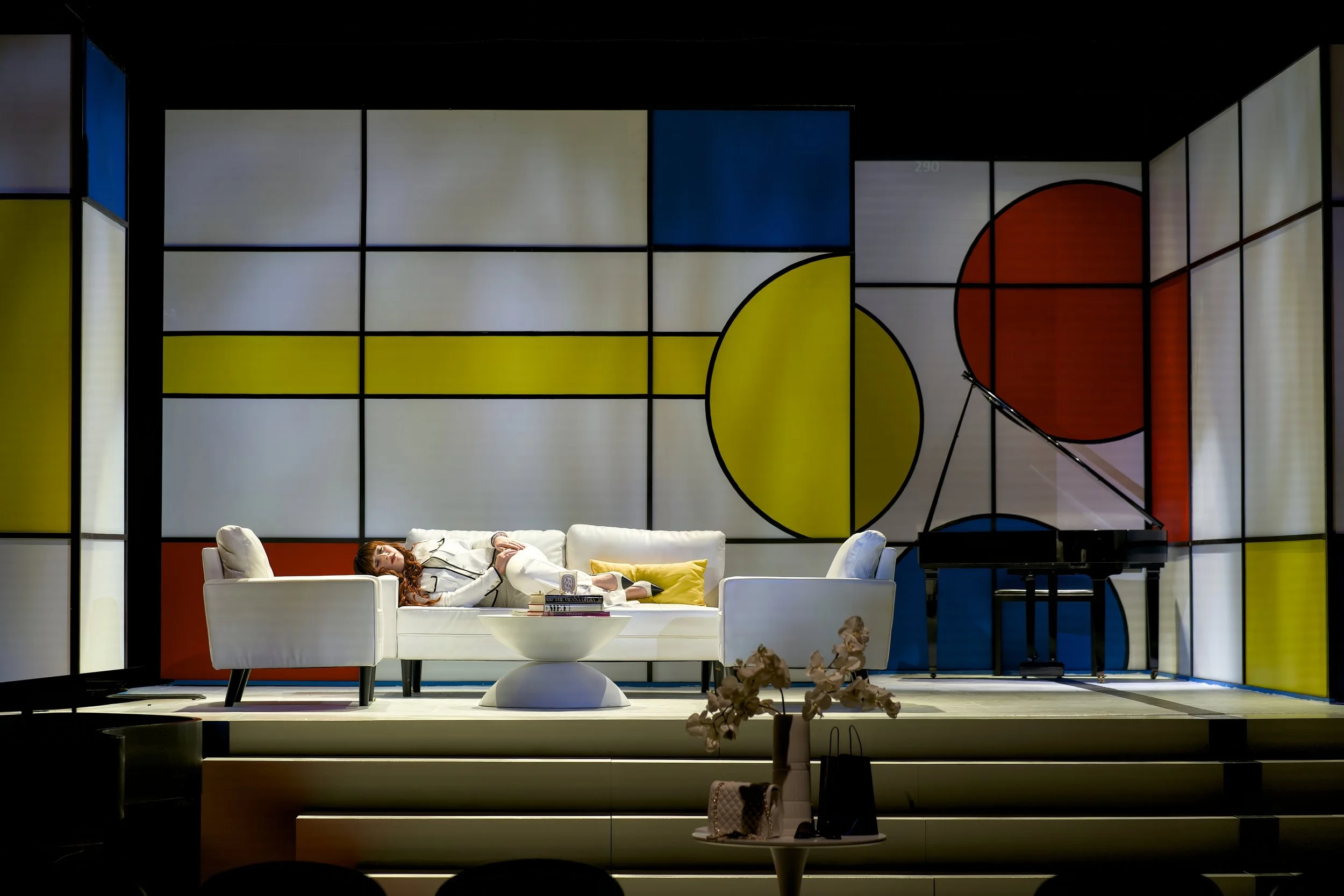 A woman dressed in white is lying on a white sofa on a stage with a colorful geometric backdrop, next to a black grand piano.