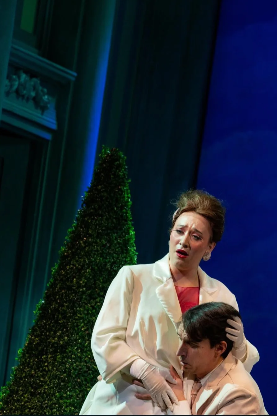 A woman in a cream-colored suit with a red dress underneath appears distressed, while a man in a light-colored suit holds her hand. There is a large green shrub or tree in the background, and the setting looks like a stage with blue and green lighting.