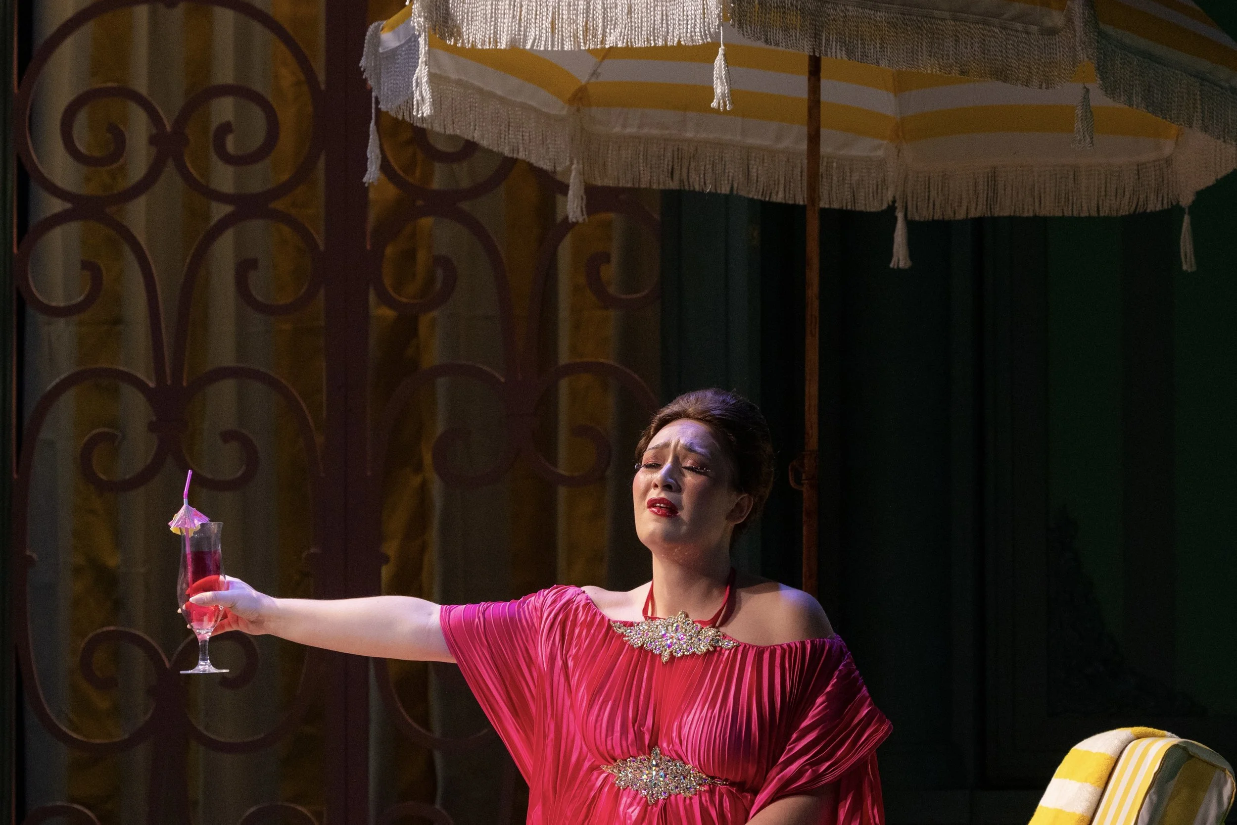 A woman in a red dress holding a glass of red cocktail with a small umbrella, standing beneath a large parasol, with an ornate metal gate in the background.