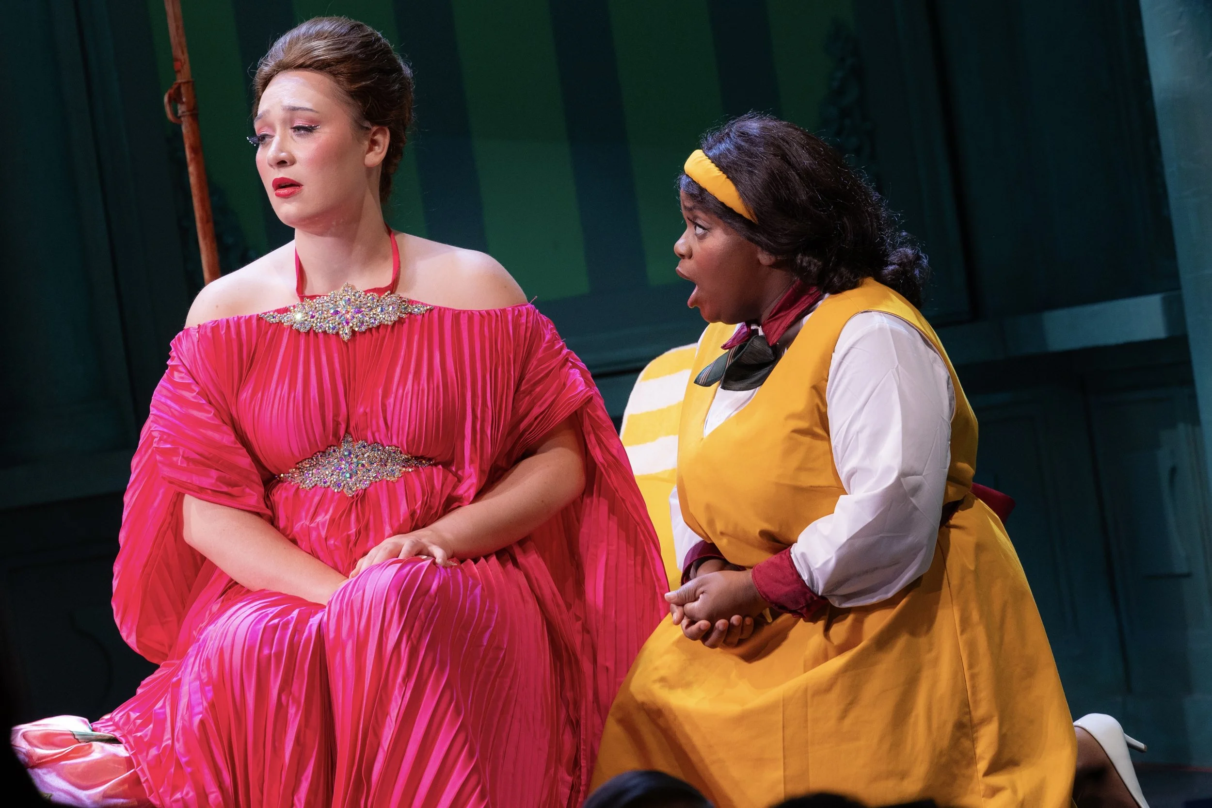 A theatrical scene with two women: one wearing a bright pink gown with off-the-shoulder sleeves and rhinestone embellishments, looking wistful, and the other in a yellow uniform with a headband, appearing to speak passionately.