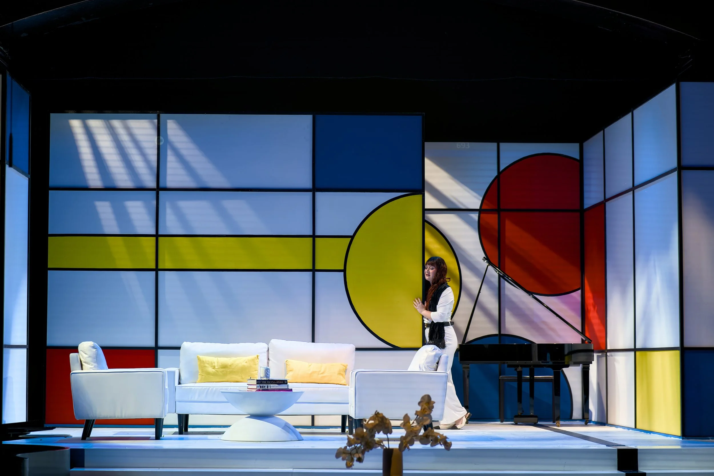 A woman standing near a white sofa with yellow pillows on a modern stage with a background featuring geometric shapes and colors, including blue, yellow, and red, with a black grand piano.