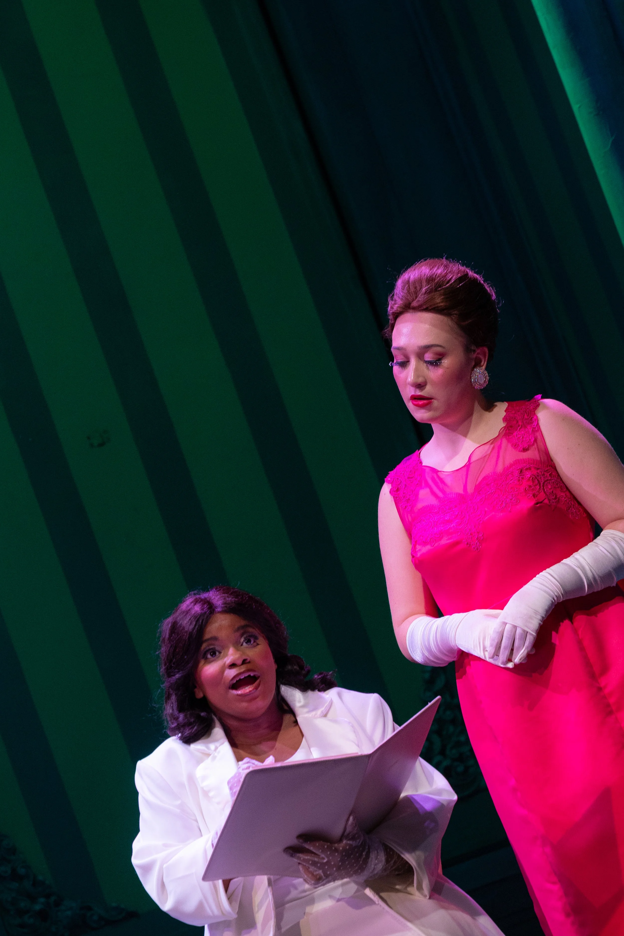 Two women on stage having a conversation, one is seated holding a folder, and the other is standing wearing white gloves and a pink dress, with a green and blue striped background.