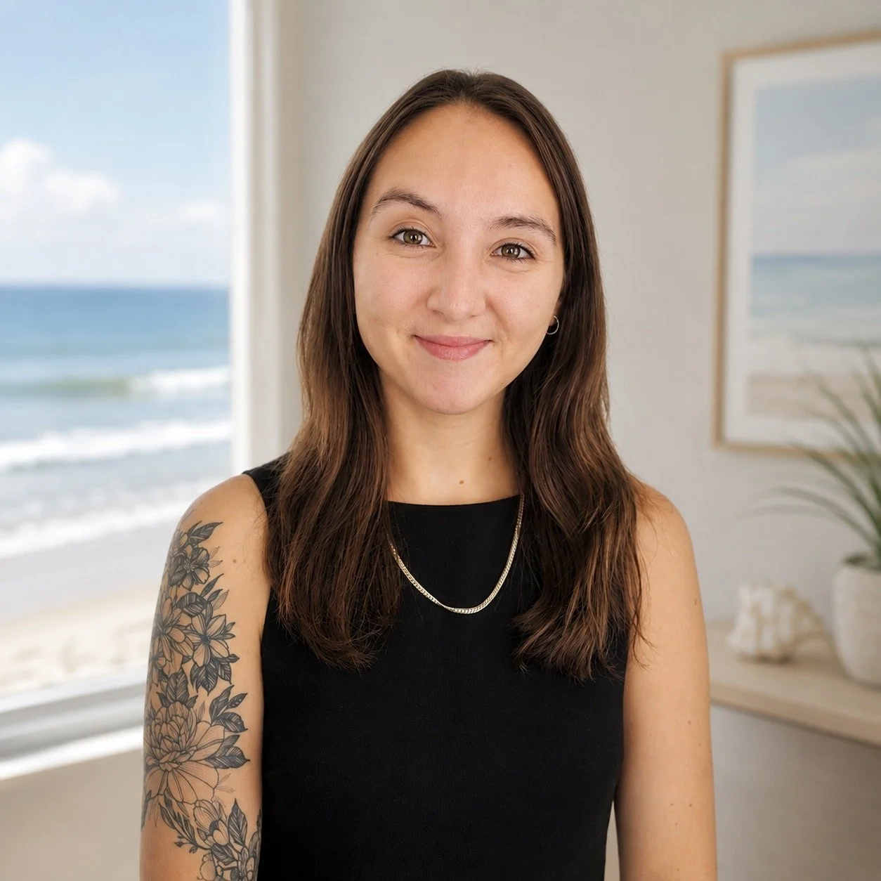 A woman with long brown hair and a floral tattoo on her left arm, wearing a black sleeveless top, standing indoors near a window with a beach and ocean visible in the background.