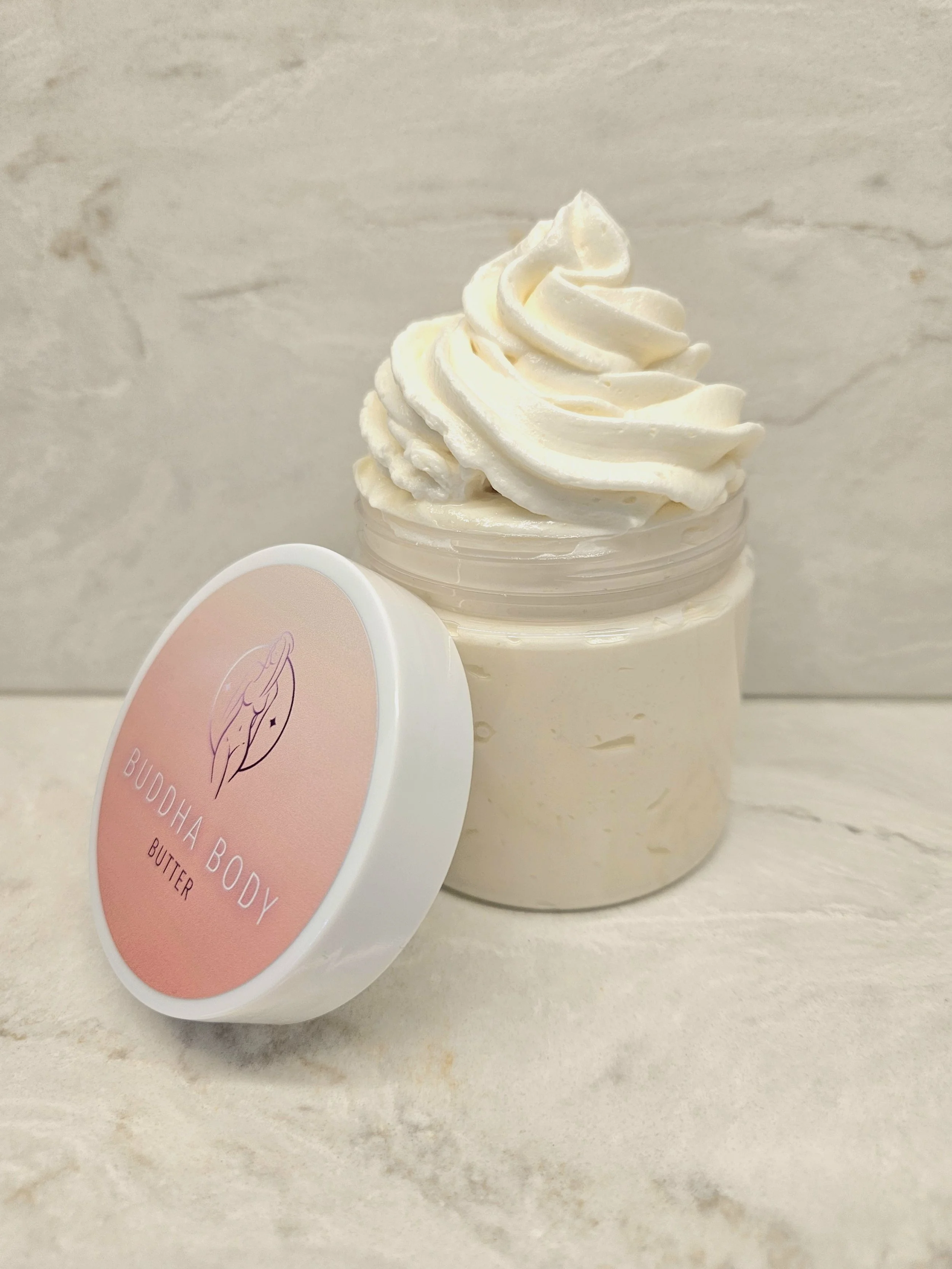 A jar of Buddha Body Butter with a swirl of whipped butter on top, placed on a light-colored surface with a textured, light-colored background.