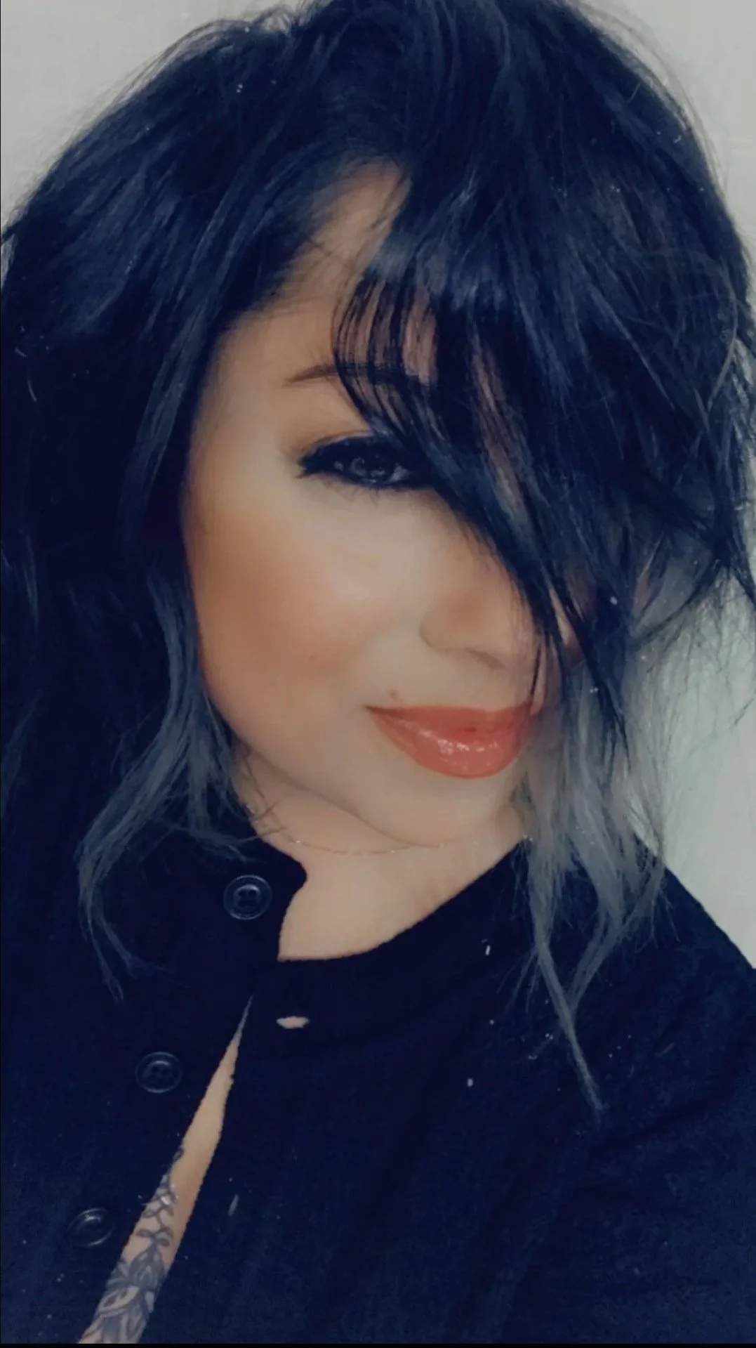 A woman with dark, wavy hair partially covering her face, smiling with red lipstick, wearing a black buttoned top.
