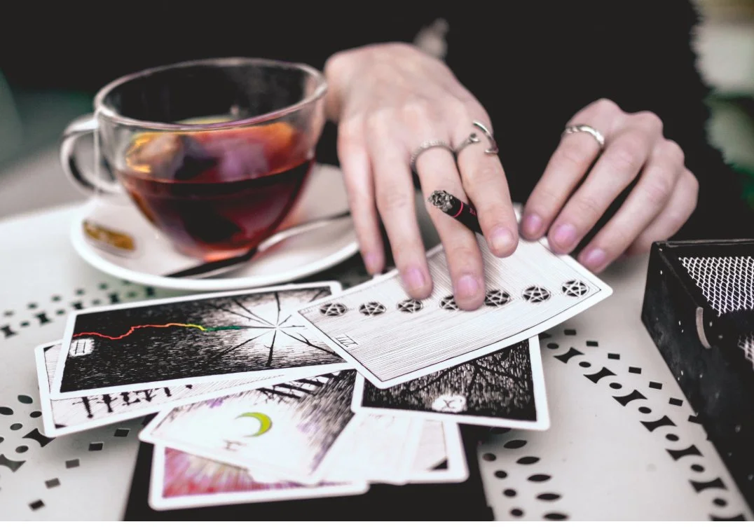 Person with multiple rings on fingers playing with tarot cards on table, with a cup of tea or coffee in the background.