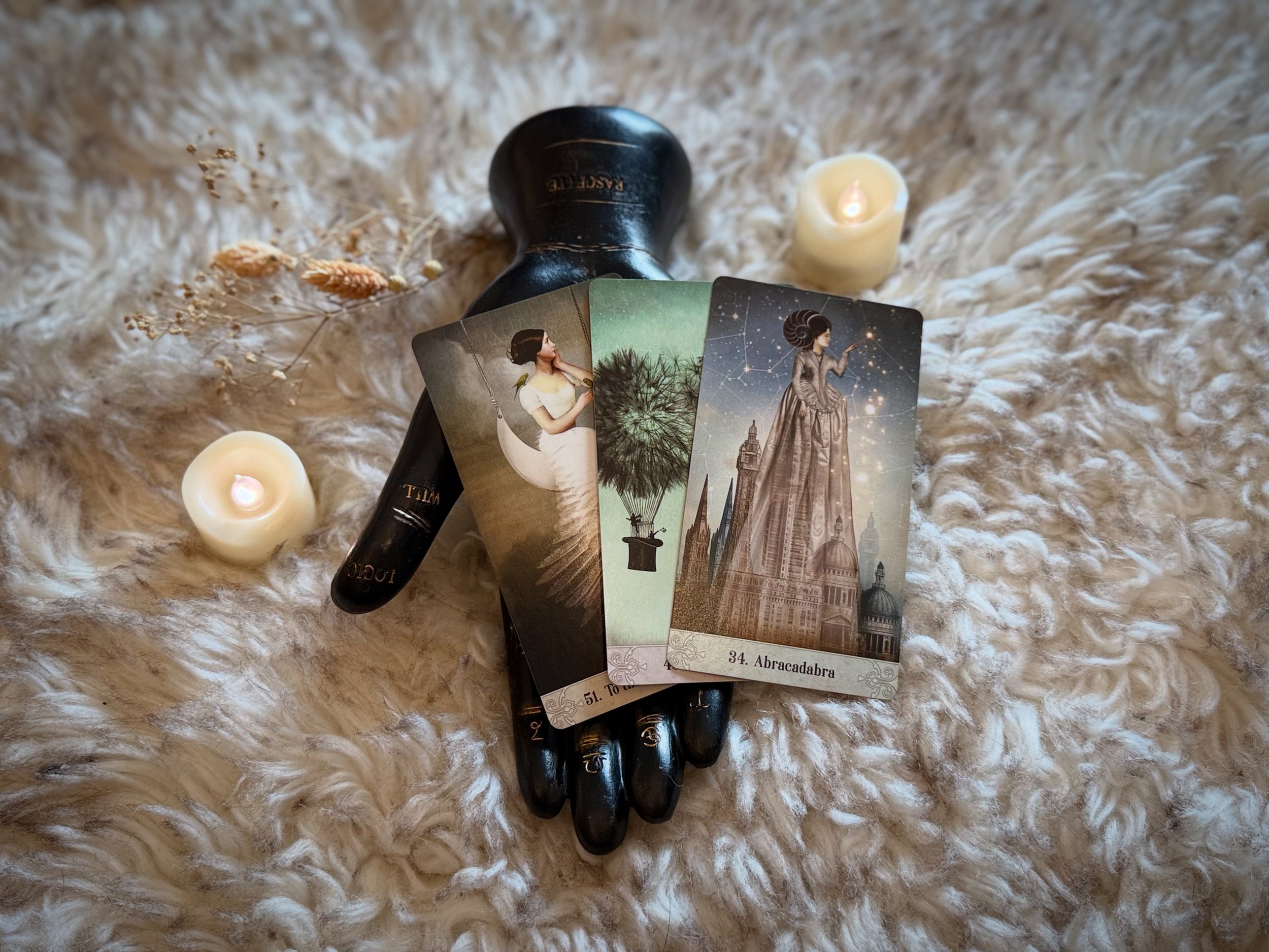 A mystical arrangement on a soft, fluffy surface featuring three tarot cards, two small lit candles, and dried decorative plants. The tarot cards depict a woman in white with wings, a potted tree with glowing leaves, and a woman in an elegant gown under a starry sky.
