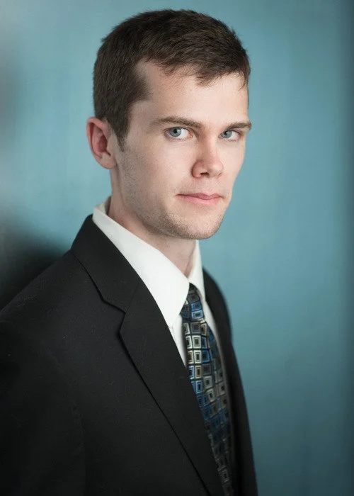 A young man with short brown hair, wearing a black suit, white shirt, and patterned tie, standing against a blue background.