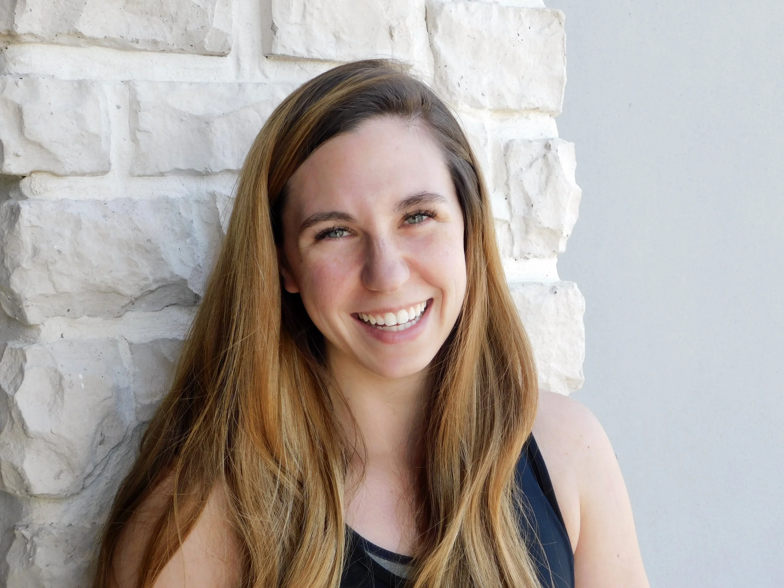 Close-up of a woman with long red hair smiling, standing in front of a white stone wall.
