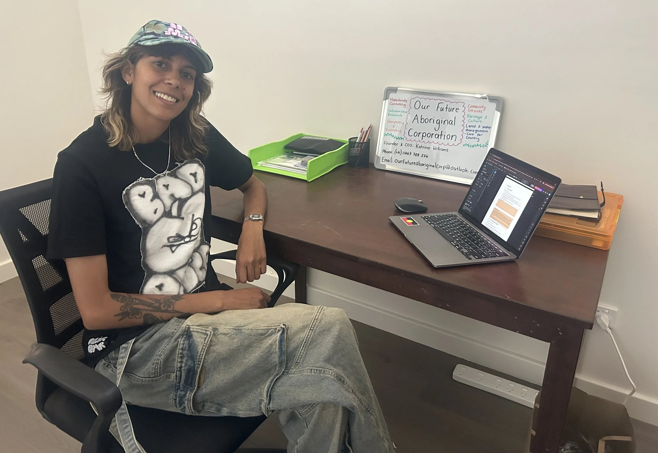 A woman sitting at a desk with a big smile, wearing a black T-shirt with a graphic, a colorful cap, and earphones. The desk has a laptop, a green tray holding papers, a black container with pencils, and a whiteboard with notes about an Aboriginal corporation.