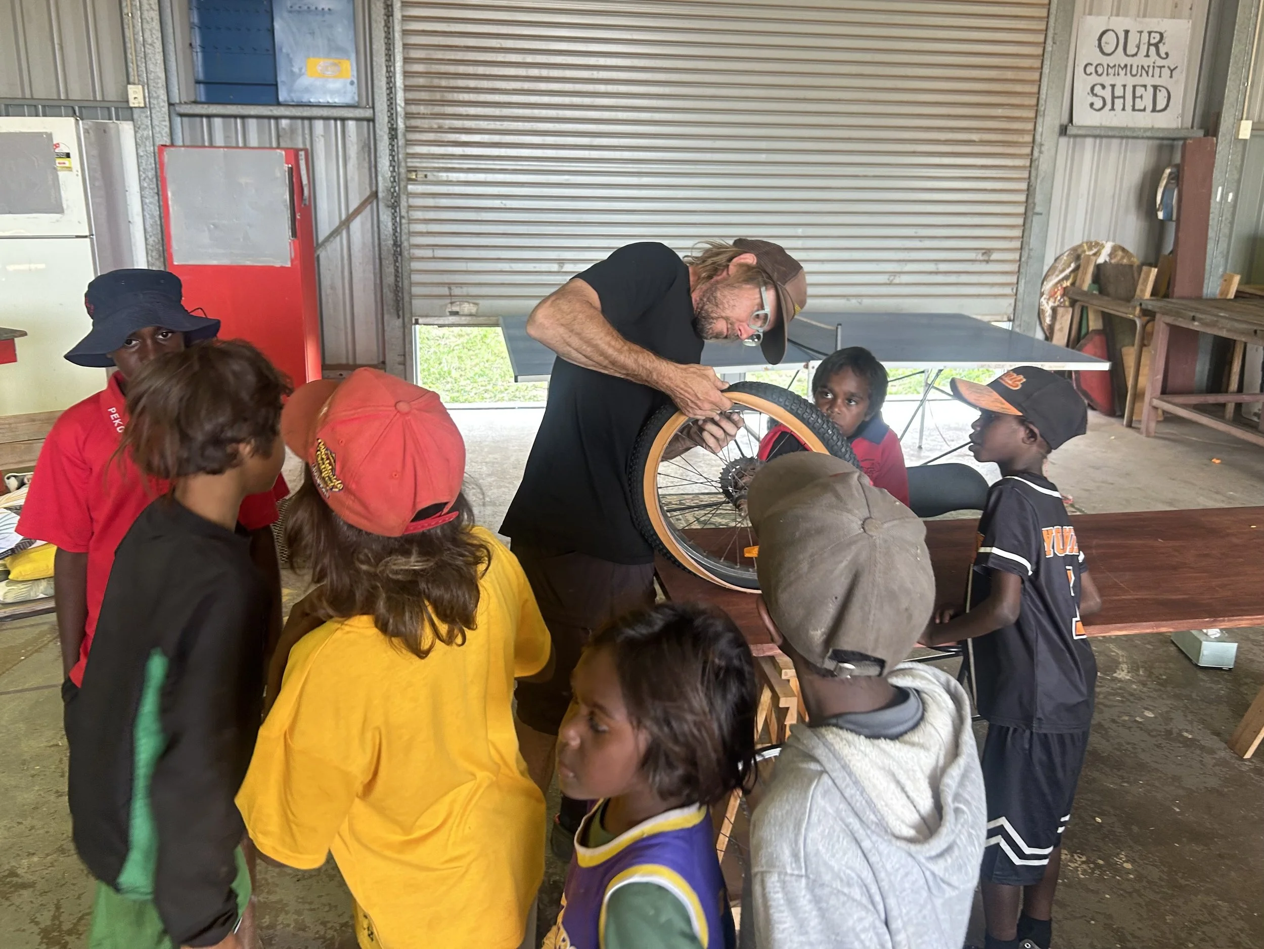 A man with glasses and long hair working on a bicycle wheel surrounded by children in a rustic shed. The shed has a sign that says "Our Community Shed" and metal walls.