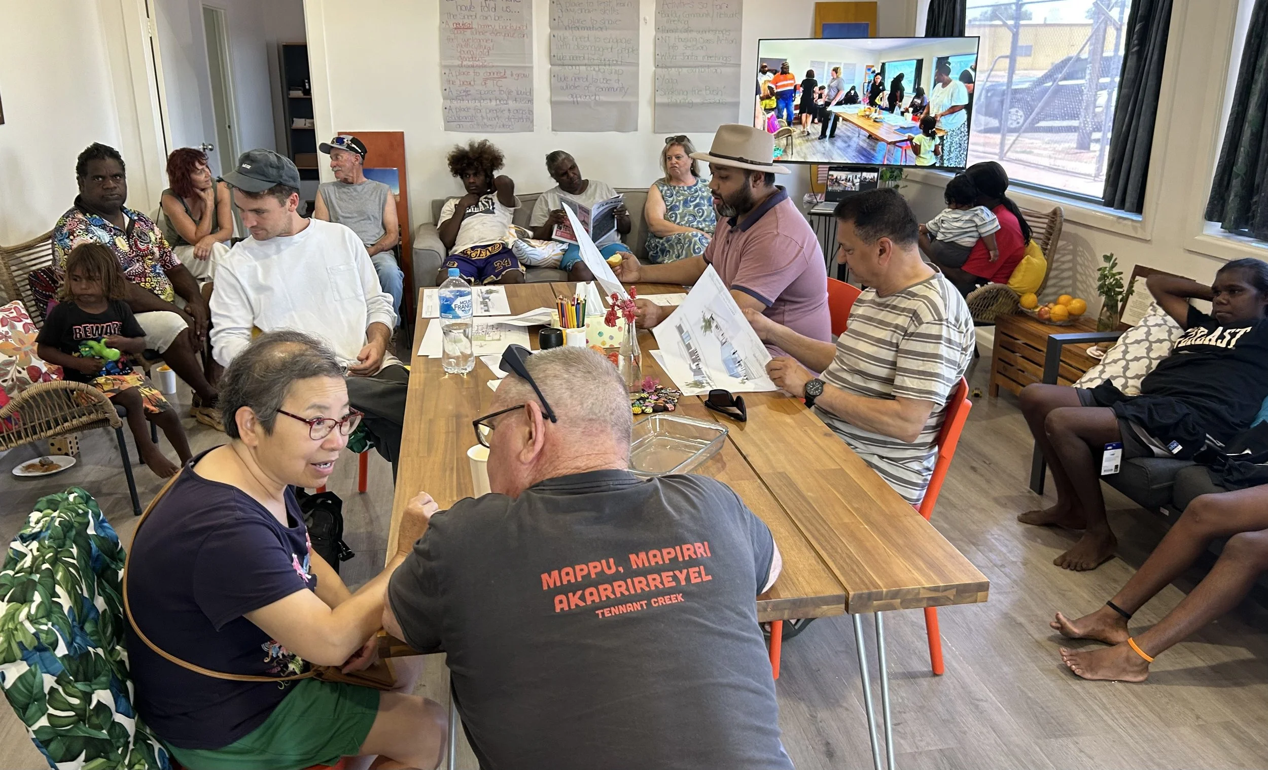 A group of diverse people gathered in a room for a meeting or workshop, with some seated at a table and others on chairs and couches. There are notes on large sheets of paper on the wall and a large screen displaying an outdoor scene of people at a community event.