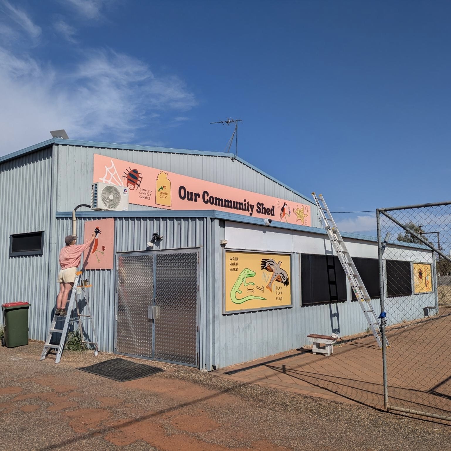 A person on a ladder painting a wall in a community shed with colorful animal and dinosaur artwork, a large sign reading 'Our Community Shed,' and a chain-link fence in the foreground under a blue sky with some clouds.
