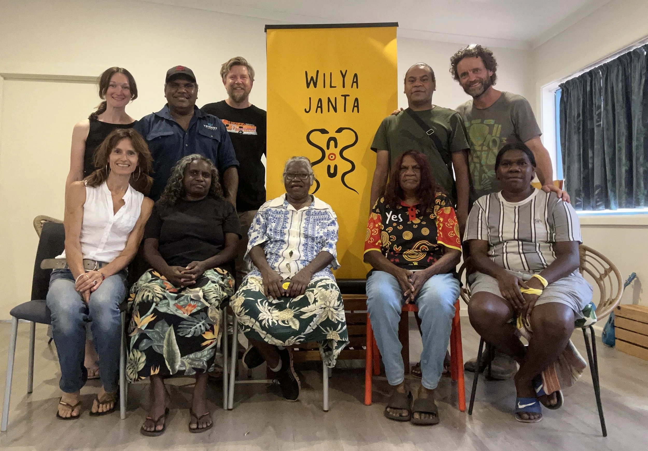 A group of nine people, six sitting and three standing, gathered in front of a yellow banner with black text and an abstract black and orange snake logo. The banner reads 'WILYA JANTA' and features a stylized snake design. The group appears to be at an indoor event or gathering, with diverse ages and ethnicities.