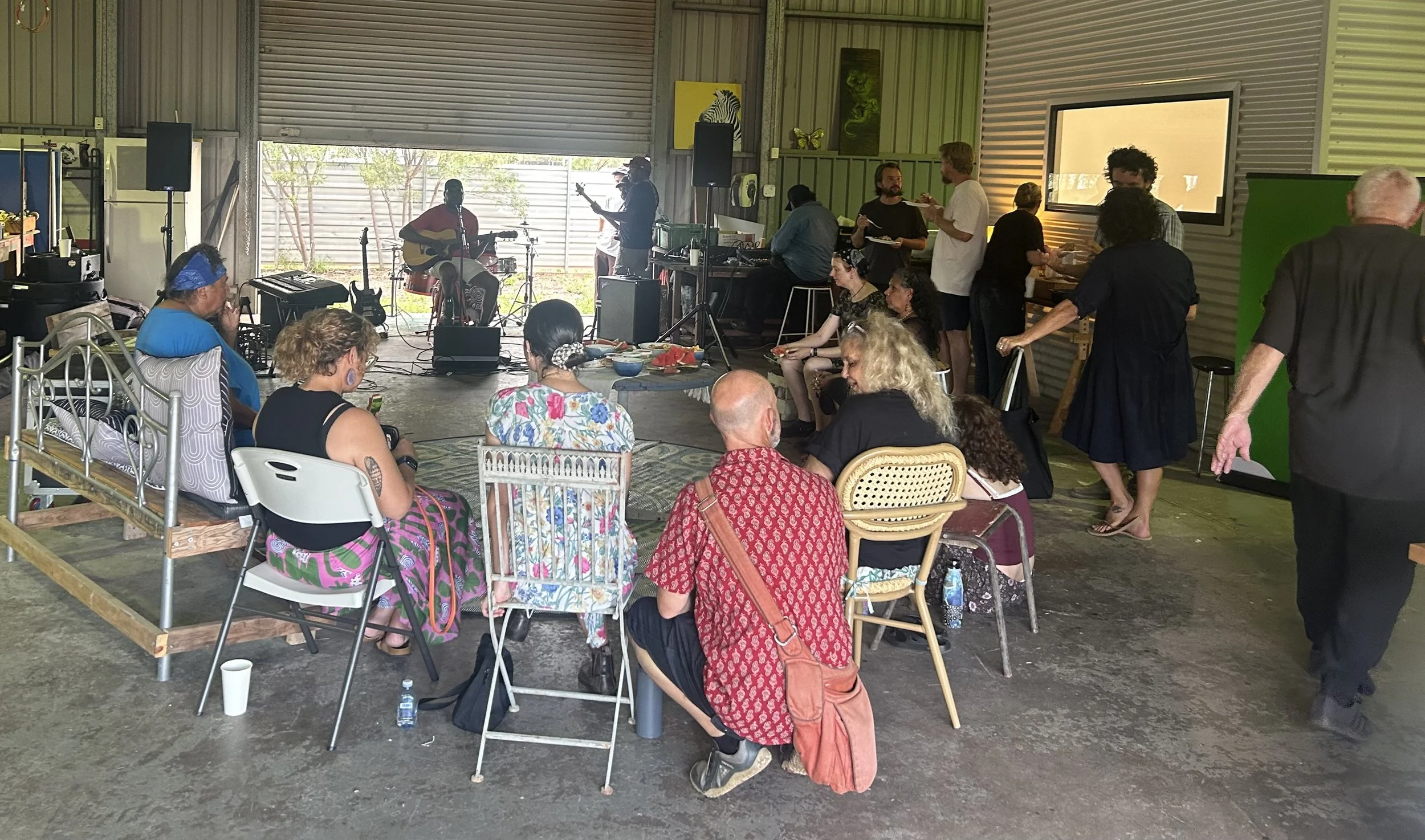 A group of people gathered indoors watching a live music performance. The band includes a guitarist and a bassist. The audience is sitting and standing, some eating, with tables of food and drinks. The space has metal walls and a large open garage door.
