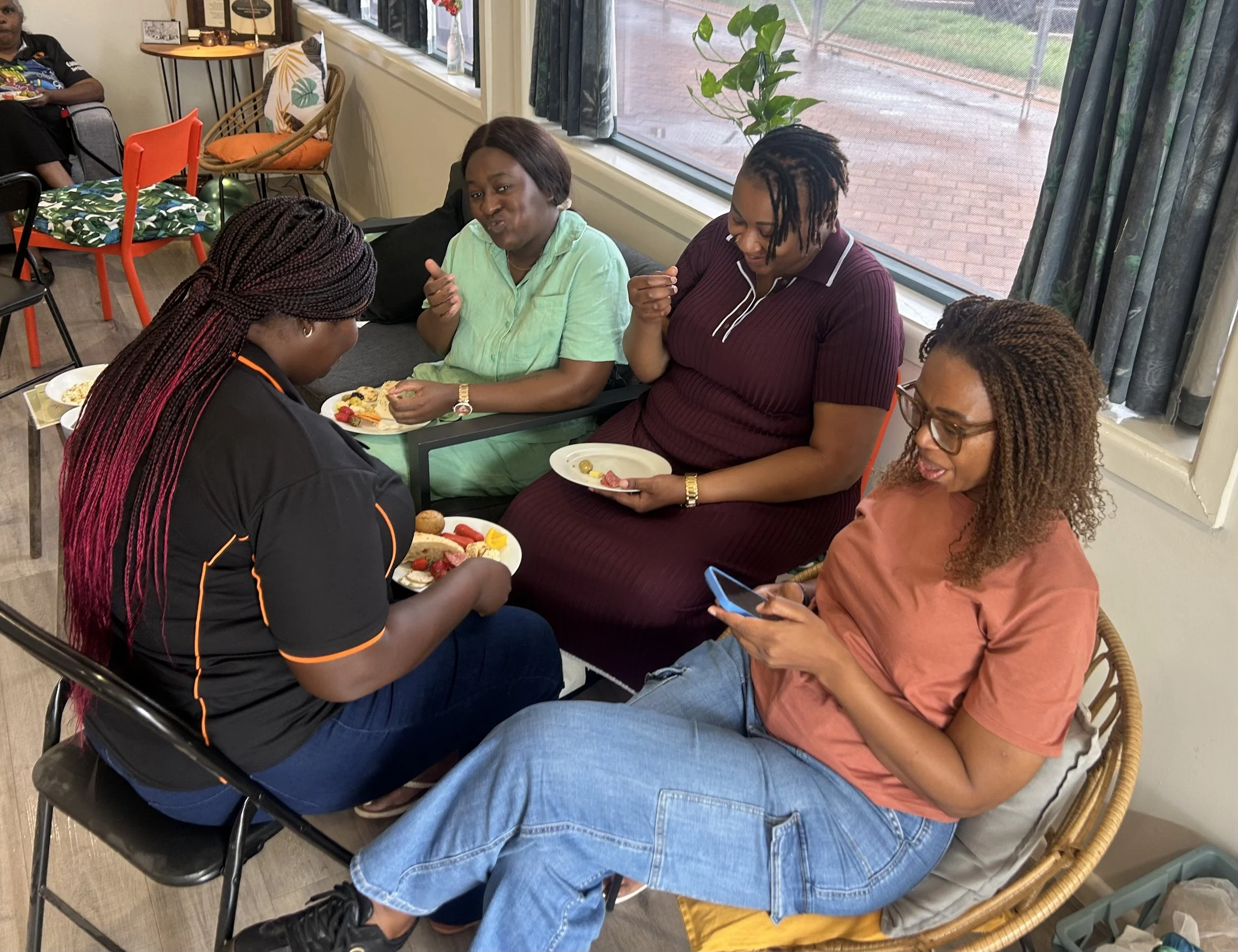 A group of four women sitting at a table in a cafe, eating and talking, with a window showing brick pavement outside.