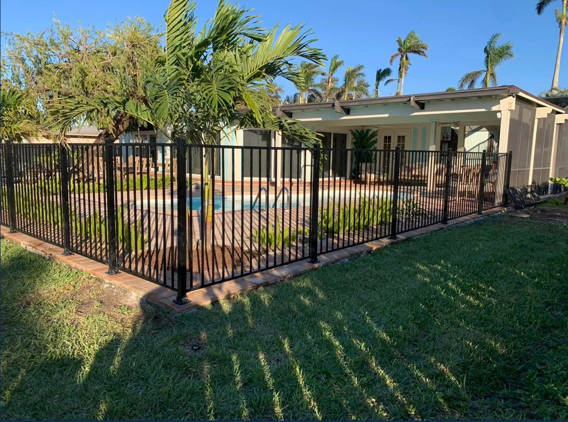A fenced backyard with a swimming pool, surrounded by green grass, tropical plants, and a house with a covered patio, under a clear blue sky.
