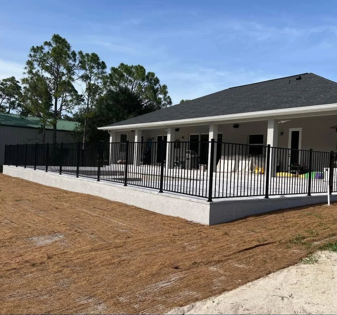 A newly constructed house with a black metal fence surrounding a concrete patio, and a yard with bare soil.