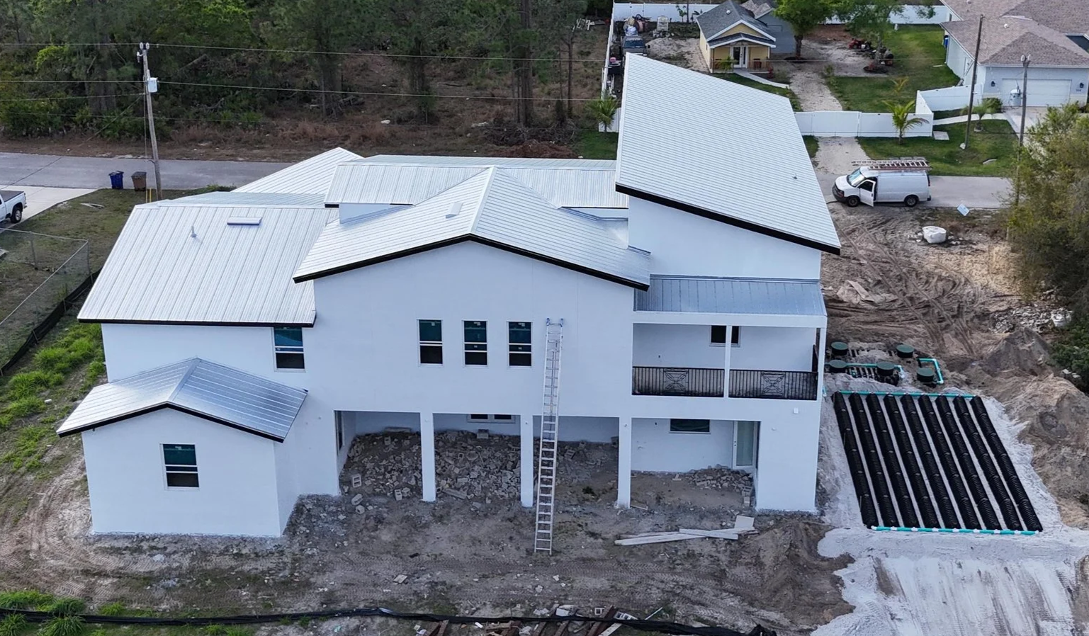 A multi-story house under construction with metal roofing, surrounded by dirt and construction materials, in a suburban neighborhood.
