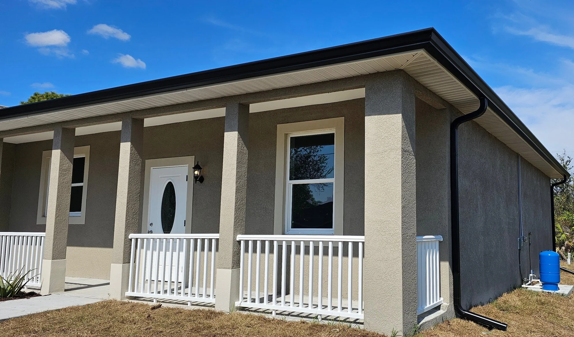 Front view of a modern house with a front porch, white railing, gray exterior walls, and a gable roof under a blue sky with clouds.