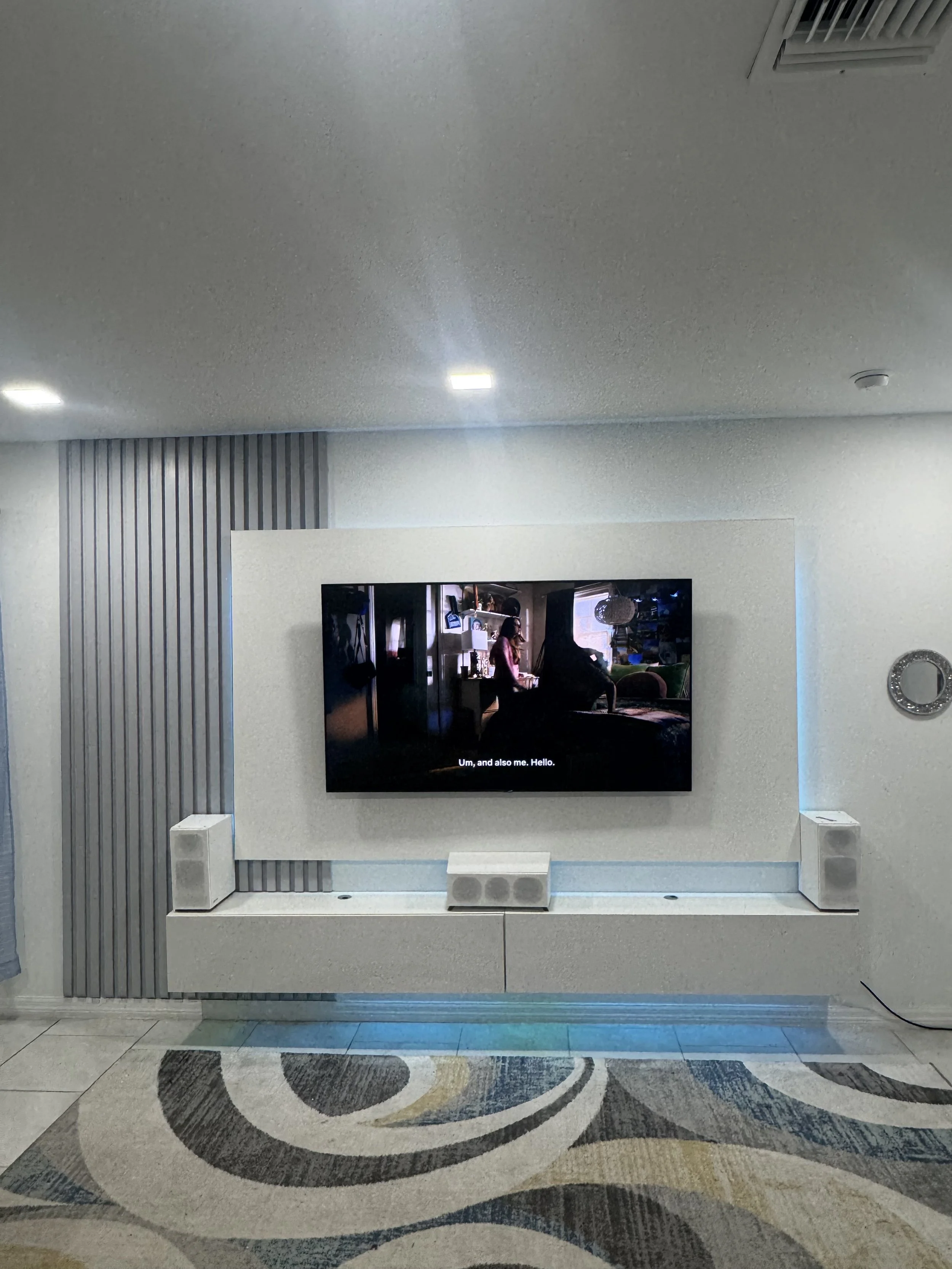 Living room with mounted flat-screen TV, white soundbar below, two white speakers on either side, gray textured wall behind, patterned area rug on tiled floor.