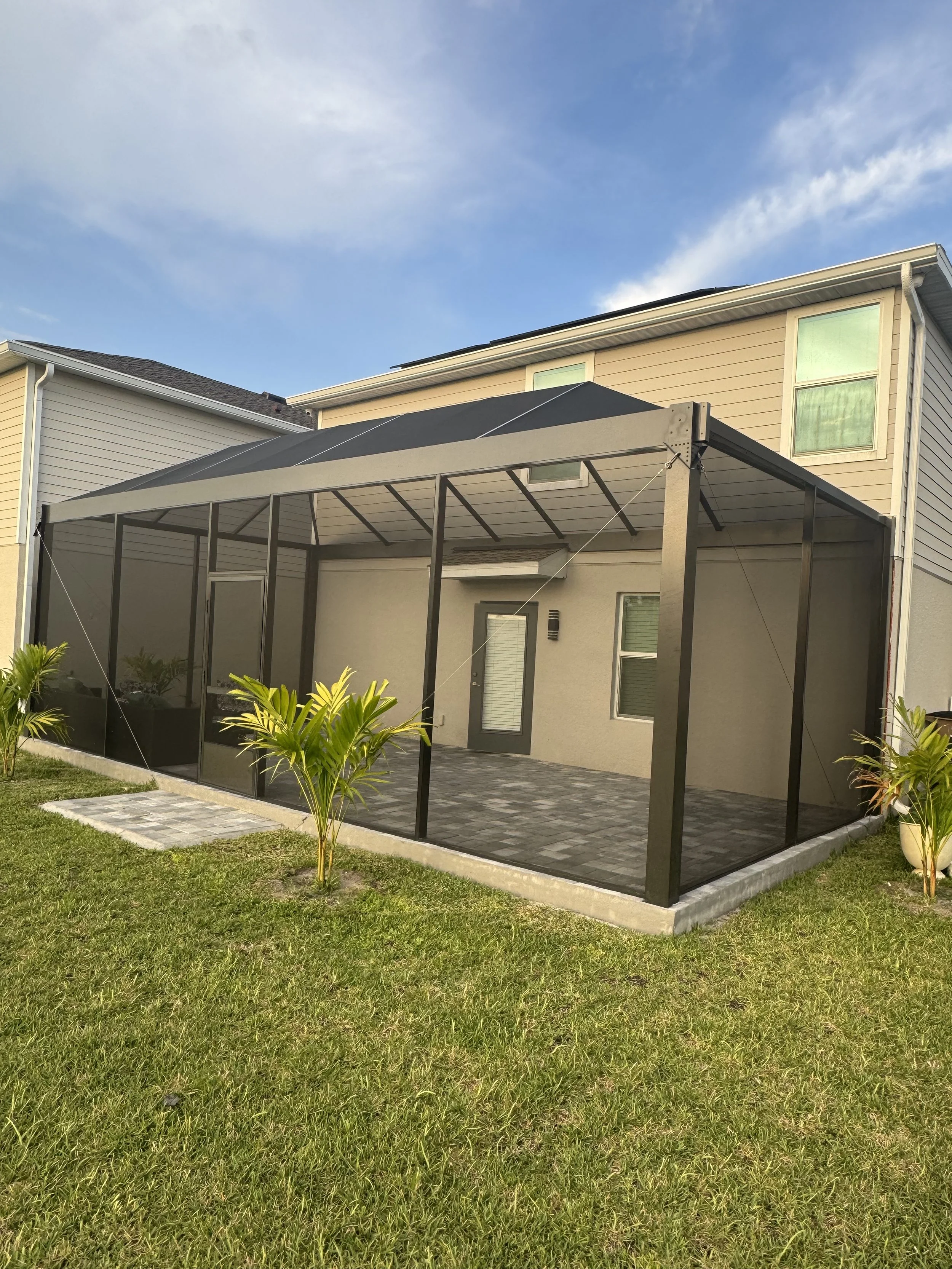 A screened-in backyard patio with a black frame, located behind a beige house with two upper windows. The patio has a stone floor and is surrounded by a grassy yard with plants, under a partly cloudy sky.