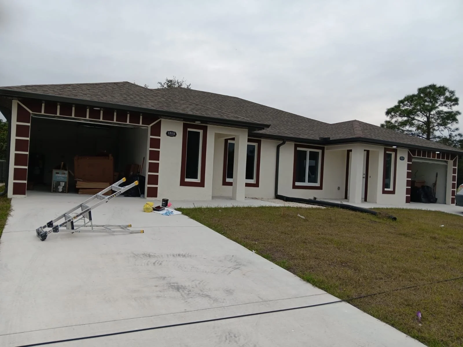 A newly constructed house with a two-car garage, white exterior with red accents, and brown shingle roof. The garage doors are open, with construction tools and materials inside. The front yard has some grass and a concrete driveway, with a ladder and construction supplies on the ground. Overcast skies overhead.