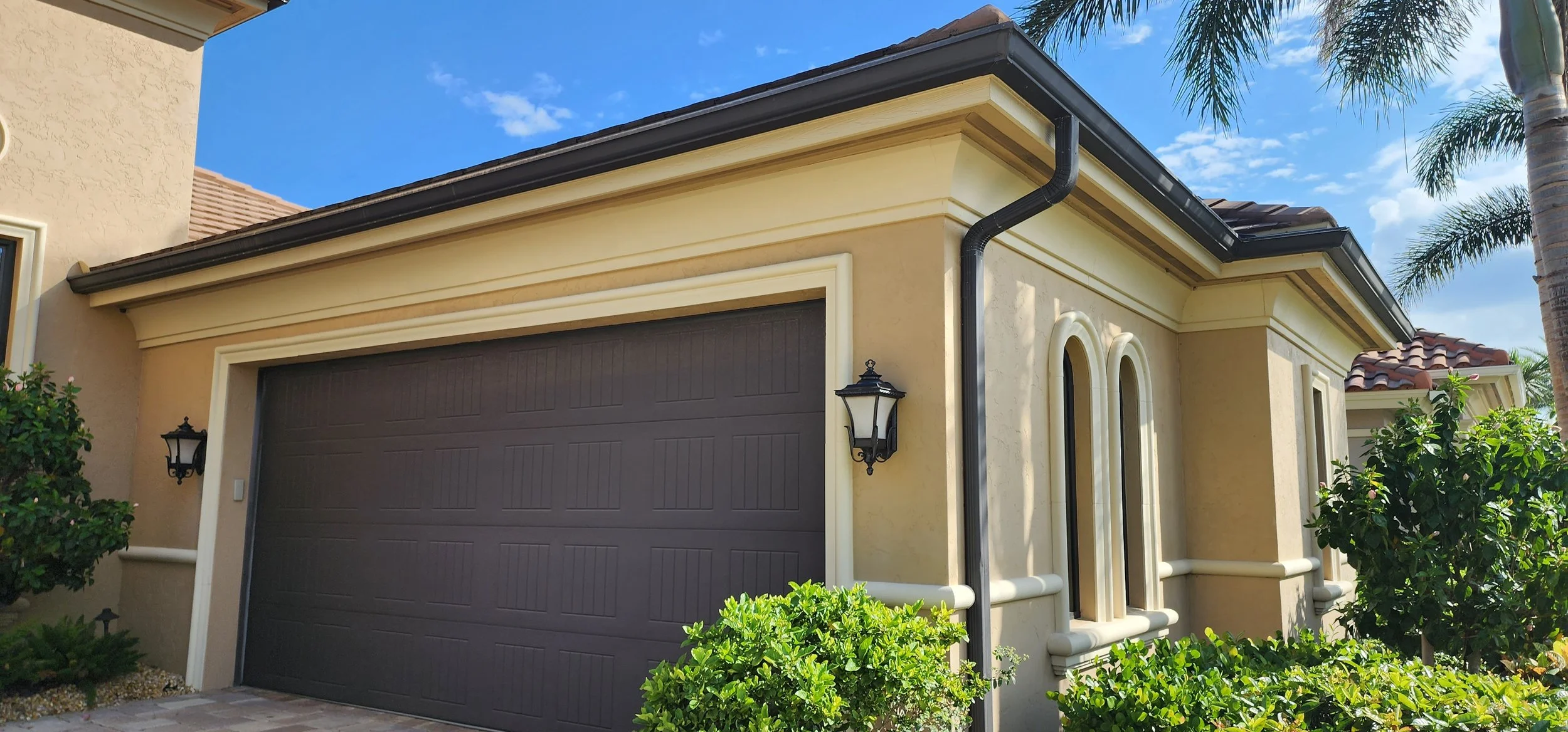 Exterior of a beige house with a brown garage door, decorative windows, black lantern-style light fixtures, green bushes, palm trees, and a blue sky.