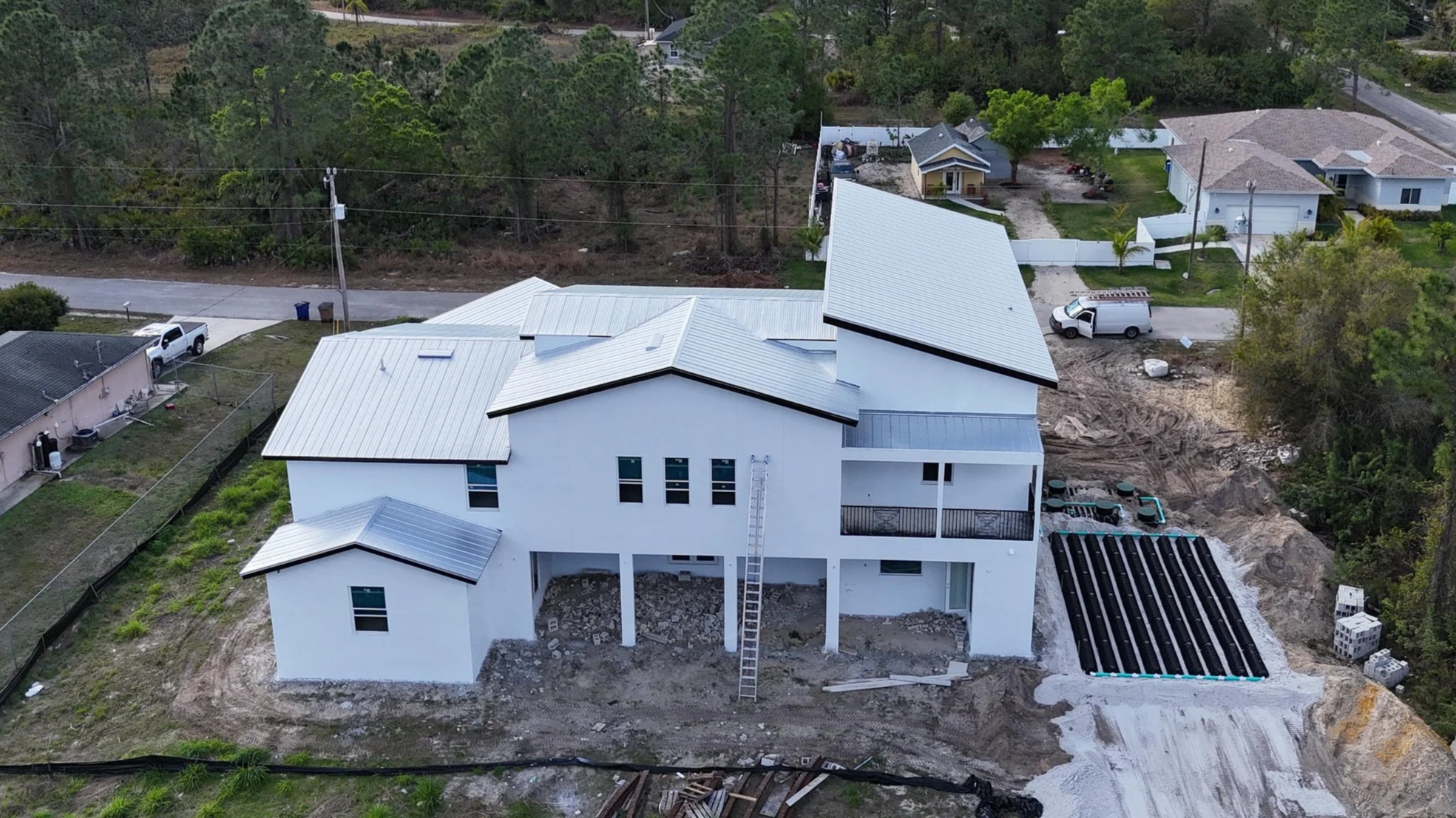 A house under construction with a metal roof in a suburban neighborhood, surrounded by trees and neighboring homes. The ground around the house shows construction debris and materials, with a construction ladder leaning against the house.