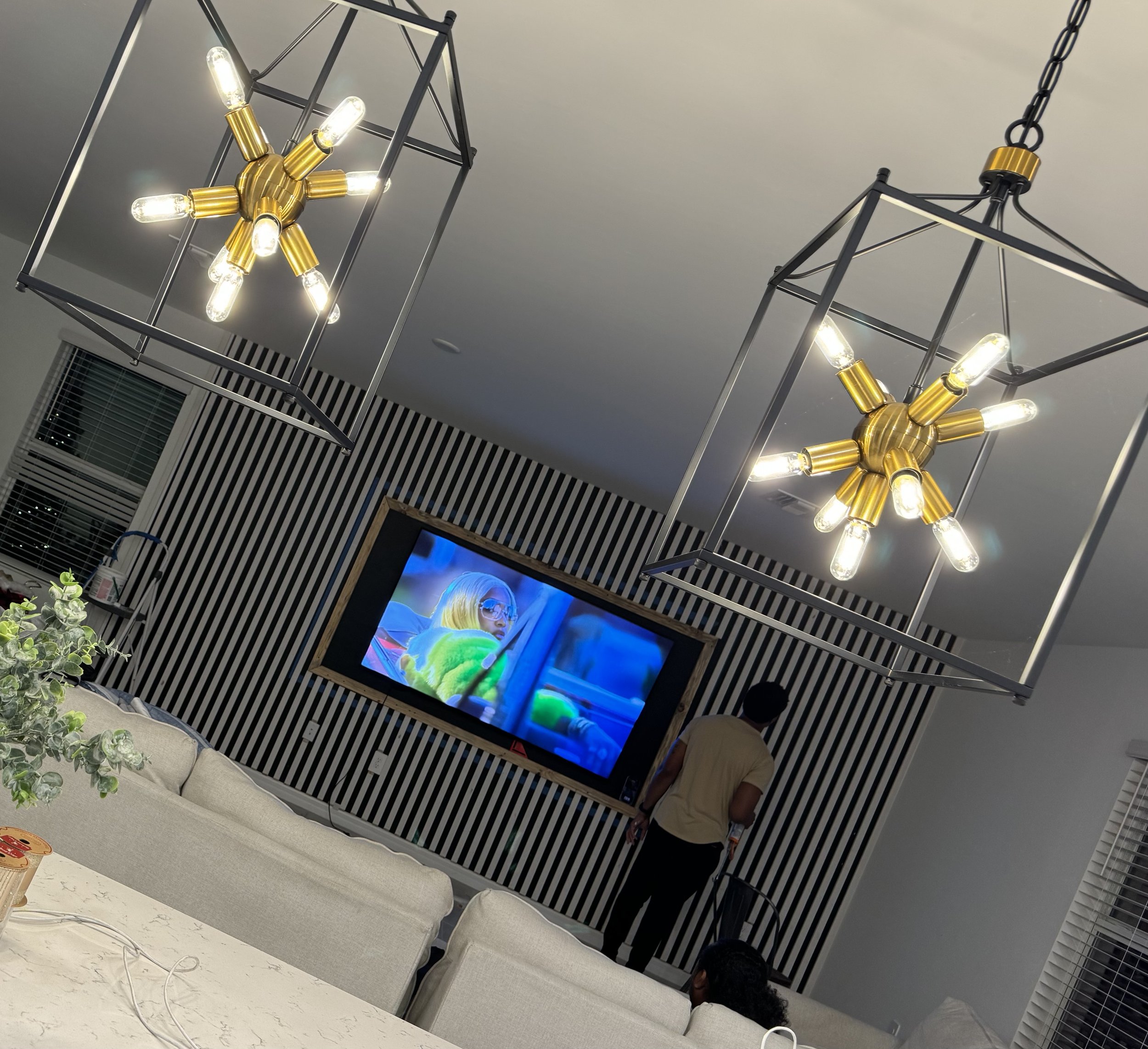 Modern living room with black and white striped accent wall, two gold and black geometric chandeliers, a wall-mounted flat screen TV showing a person in green, and a man in a beige shirt standing near the TV. There are beige couches and a white marbl