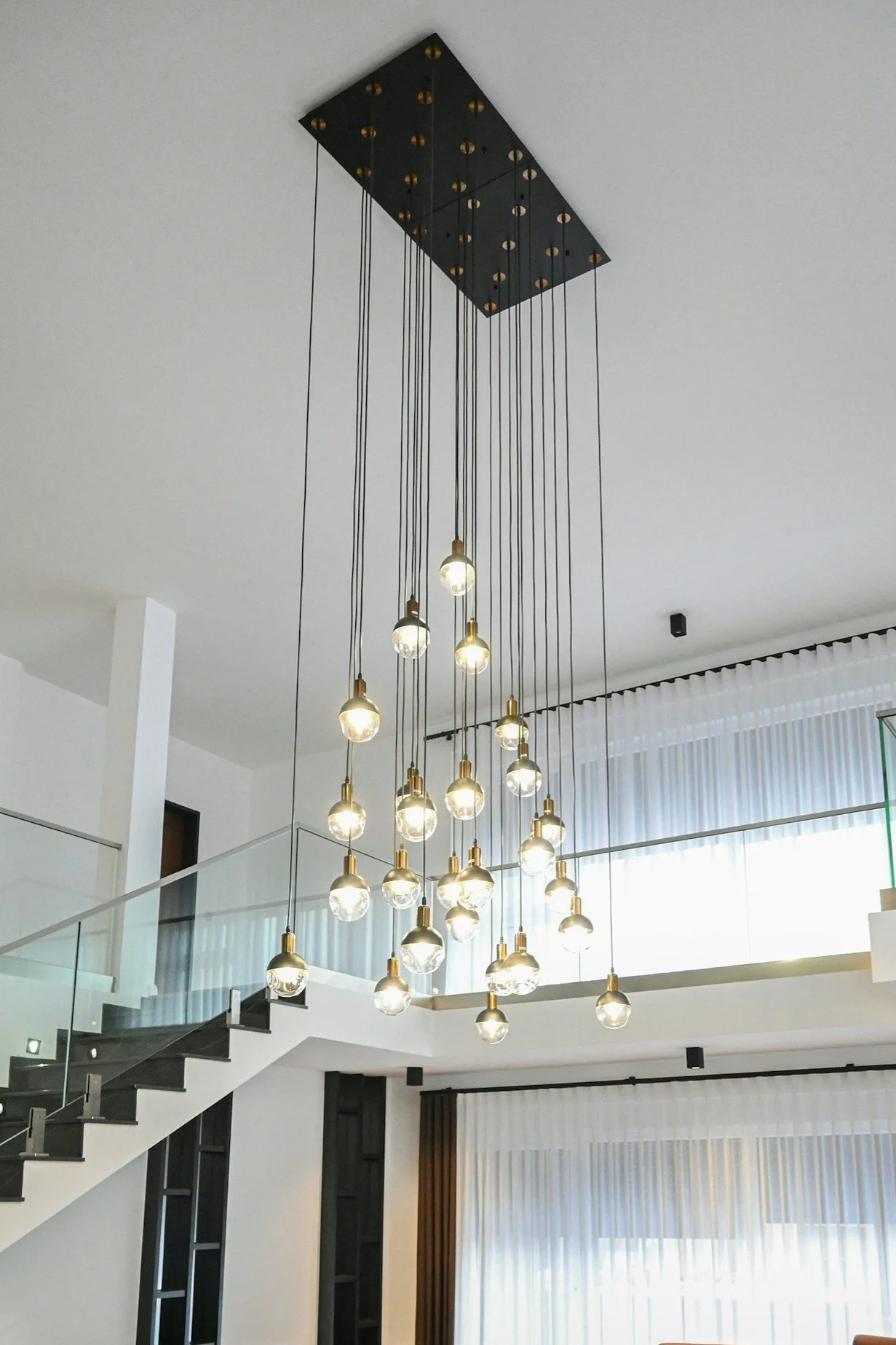Modern chandelier with multiple exposed light bulbs hanging from black cords in a spacious, bright living area with white walls, curtains, and a staircase.