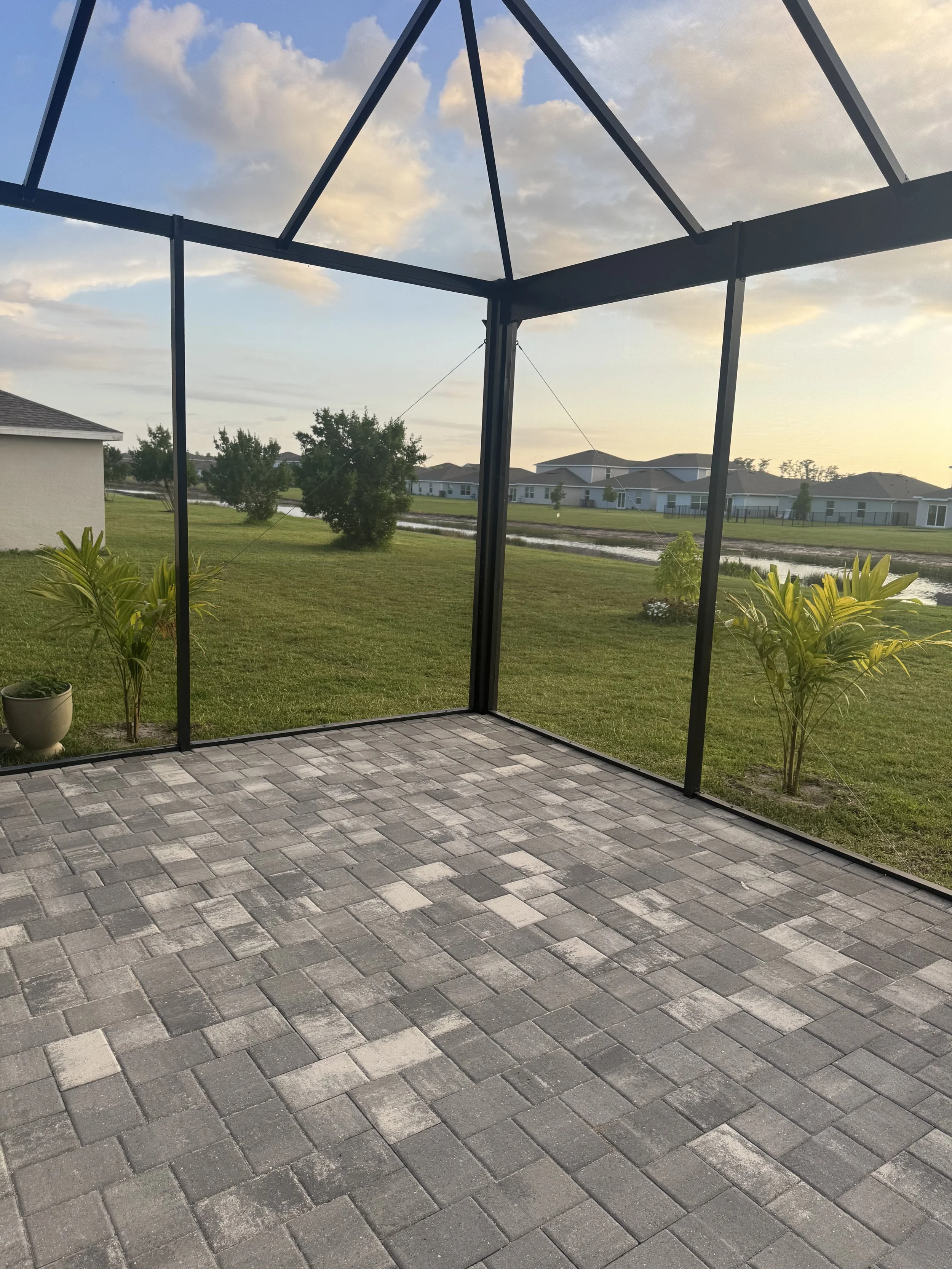 View from a screened porch with brick pavers, green grass, trees, and a neighborhood with houses and a pond in the background during sunset.