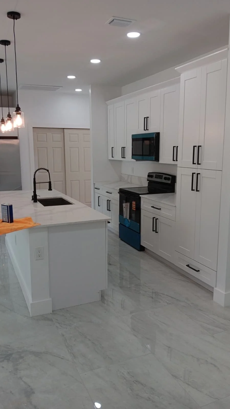 Modern white kitchen with marble floors, white cabinets with black handles, and black appliances including a stove and microwave. A kitchen island with a sink and black faucet is also visible. Hanging pendant lights are above the island.