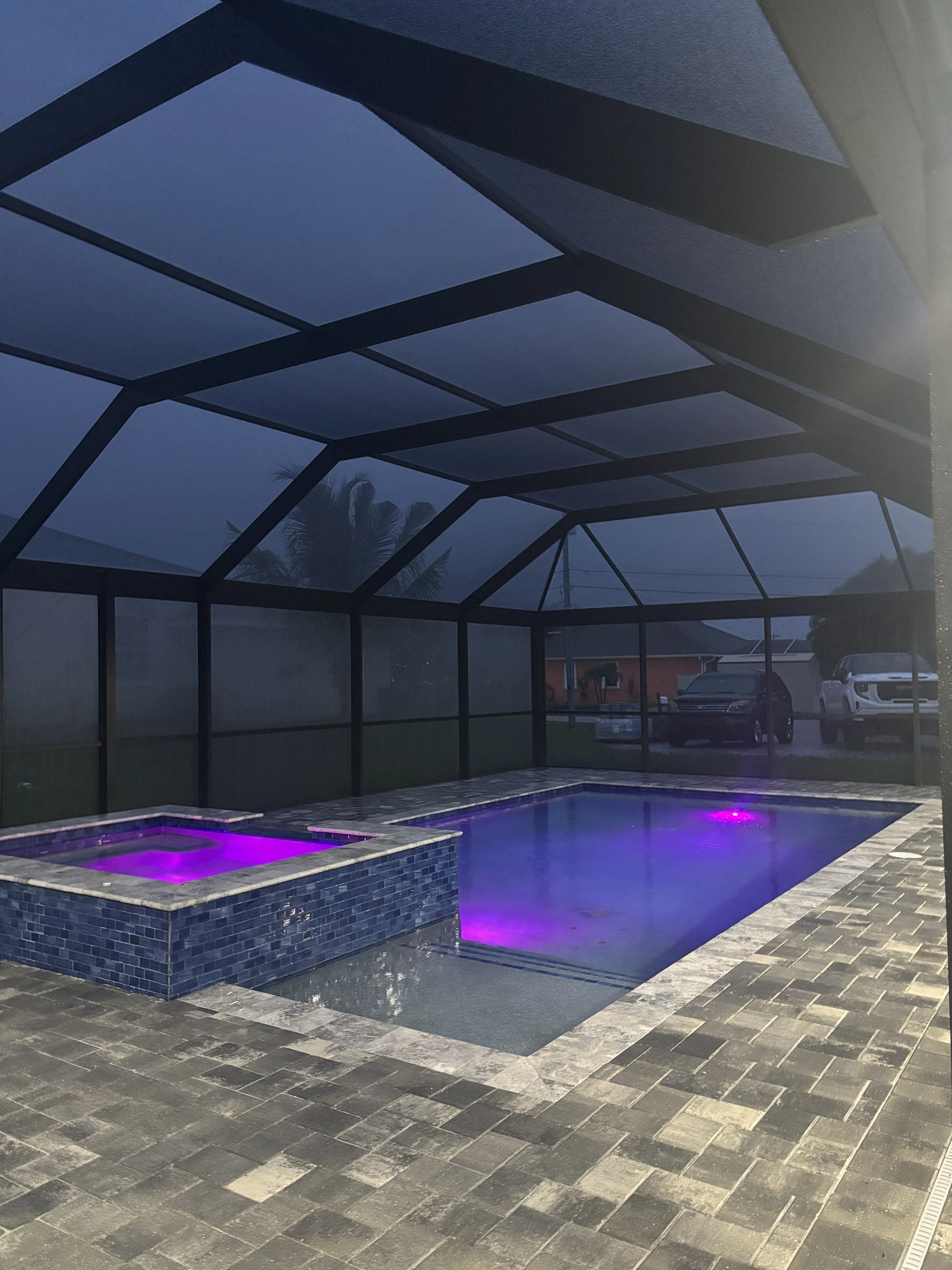 A screened-in backyard with a swimming pool and a hot tub illuminated with purple LED lights, paved with gray stone tiles, during dusk or evening.