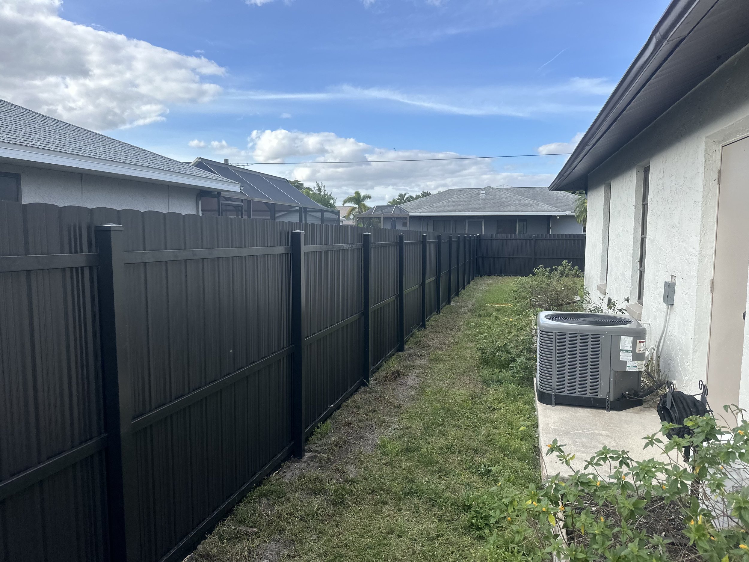 Backyard with black fence, grass, plants, and an air conditioning unit on a concrete slab, with a white house and neighboring houses in the background under a blue sky with clouds.