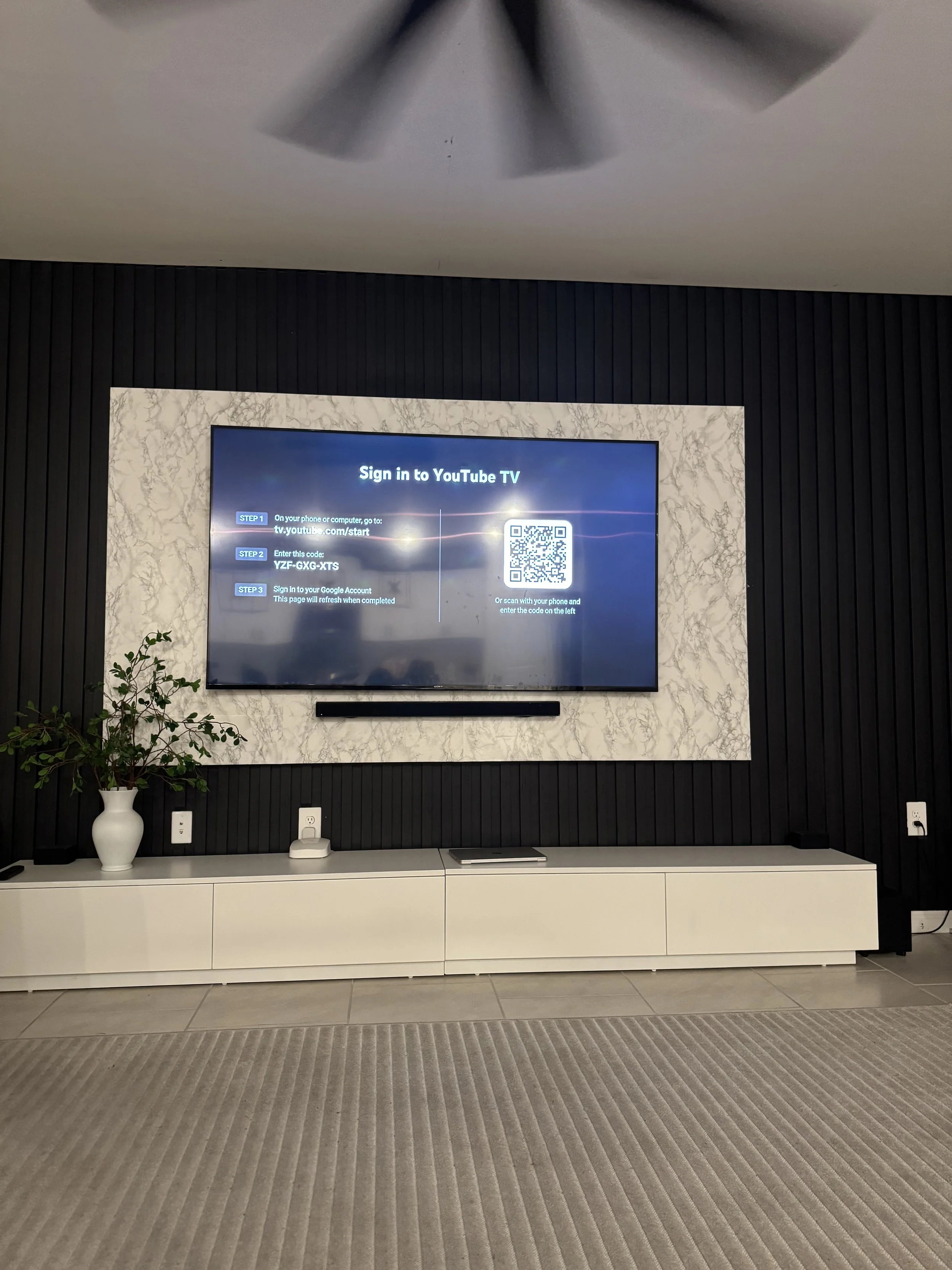 TV screen displaying instructions to sign in to YouTube TV using a QR code and a code, mounted on a marble and black wall, with a white cabinet below and a plant on the left side.