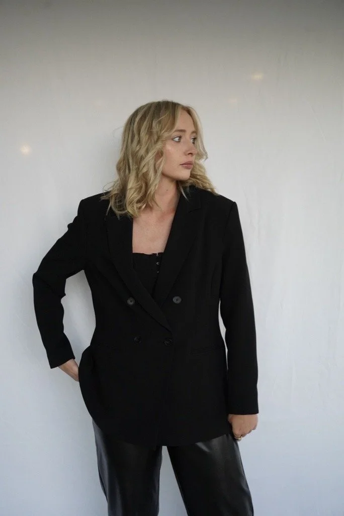A woman with blonde, wavy hair wearing a black blazer and black leather pants standing against a plain, light-colored wall.