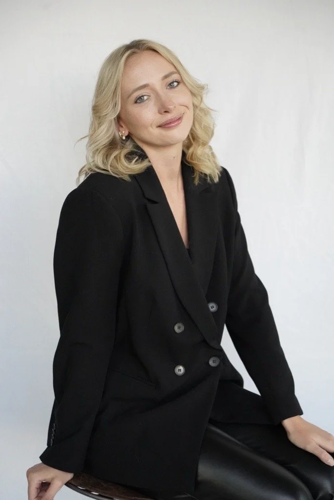 A woman with blonde hair, wearing a black blazer and black pants, sitting on a stool against a plain white background.