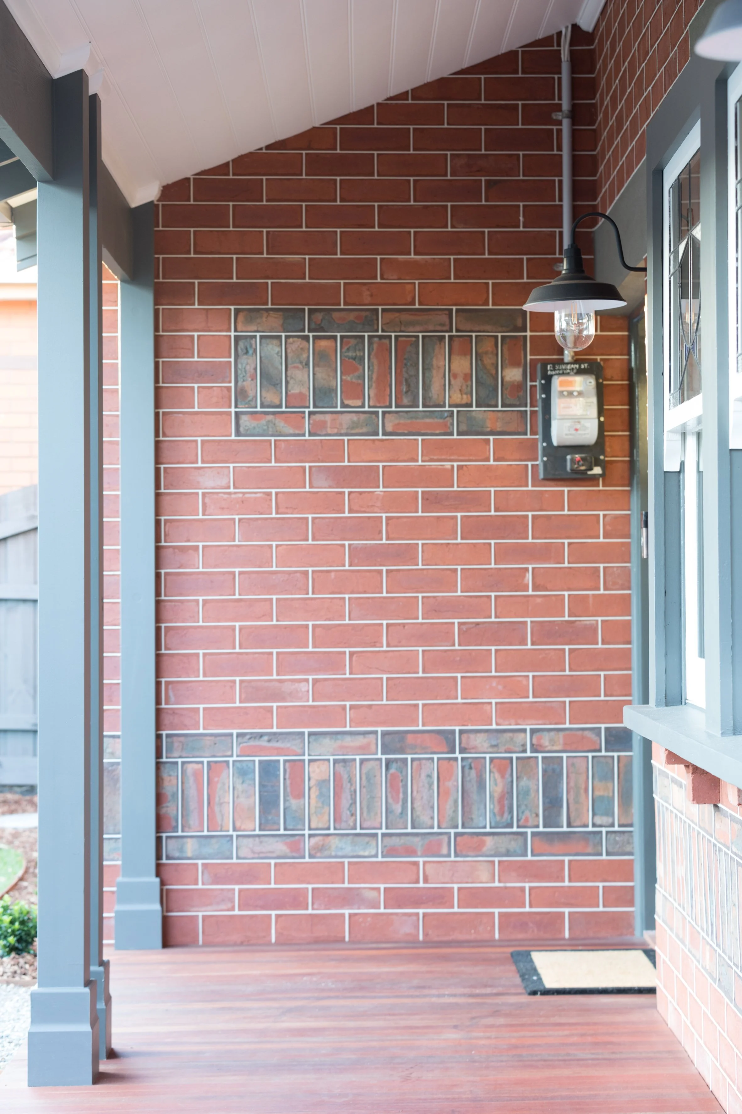 Covered porch with brick and stone wall, gray wooden pillars, a black outdoor wall light, and a beige doormat. Luxury extension and renovation, Pascoe Vale.