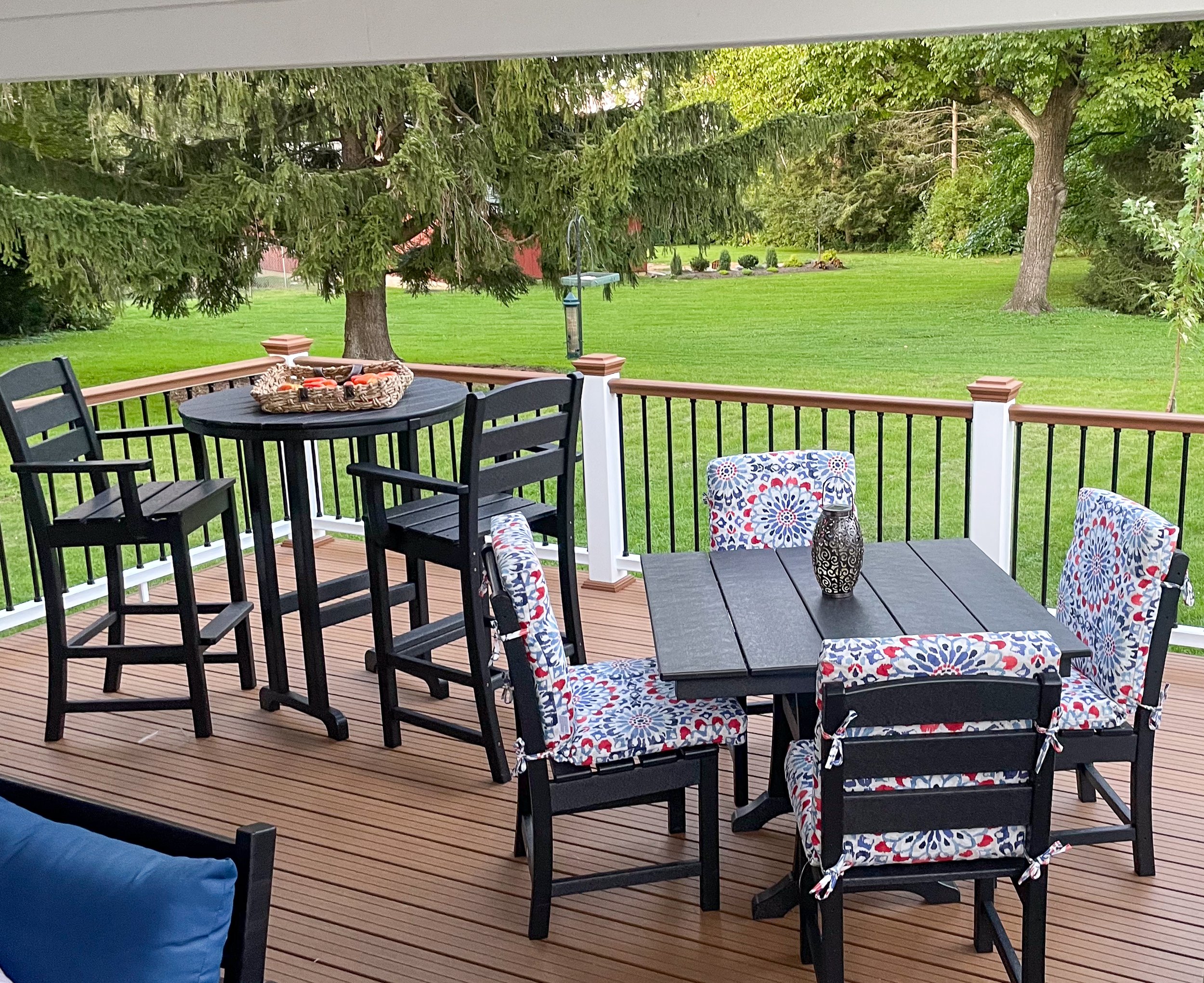 A backyard patio with black tables and chairs, some with patterned cushions, on a wooden deck surrounded by green grass and trees. There is a bird feeder hanging from a tree.