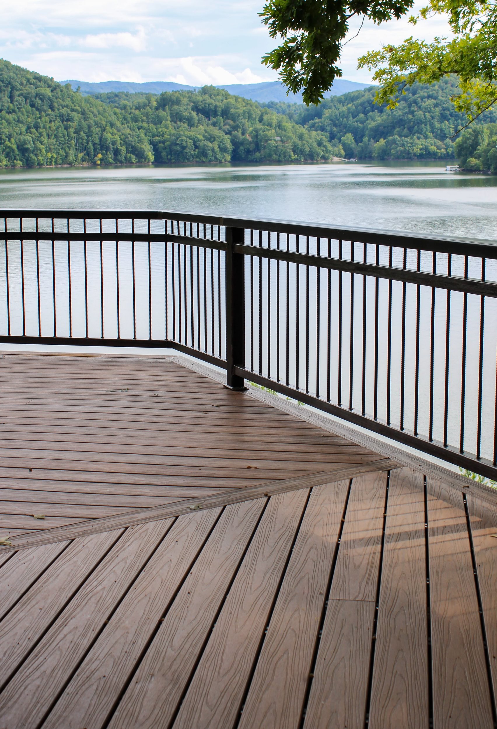 View of a expansive river surrounded by lush green mountains, seen from a wooden deck with black metal railing, under partly cloudy sky.