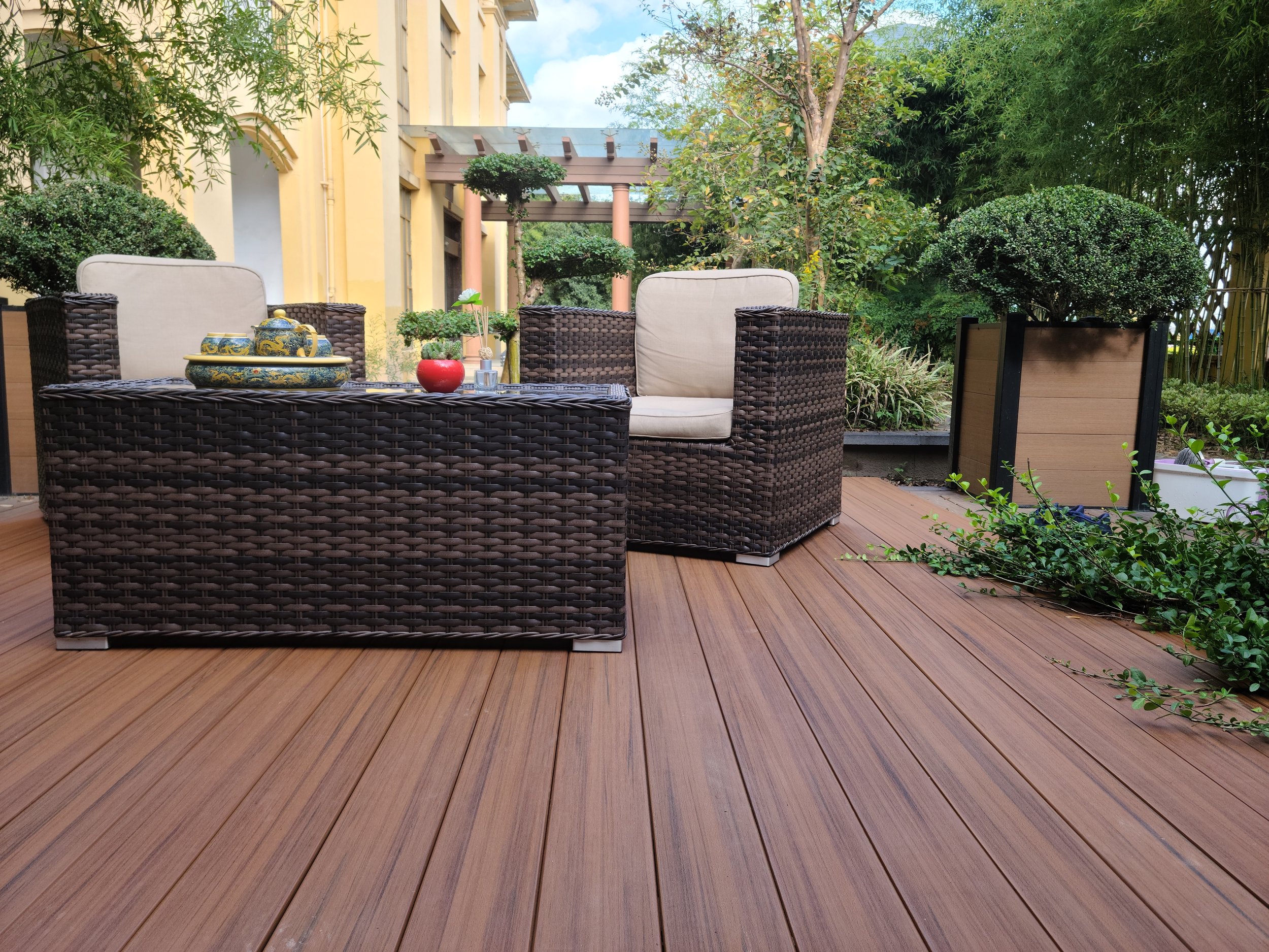 An outdoor patio area with wicker furniture, plants, and a wooden deck, adjacent to a yellow building with large windows.