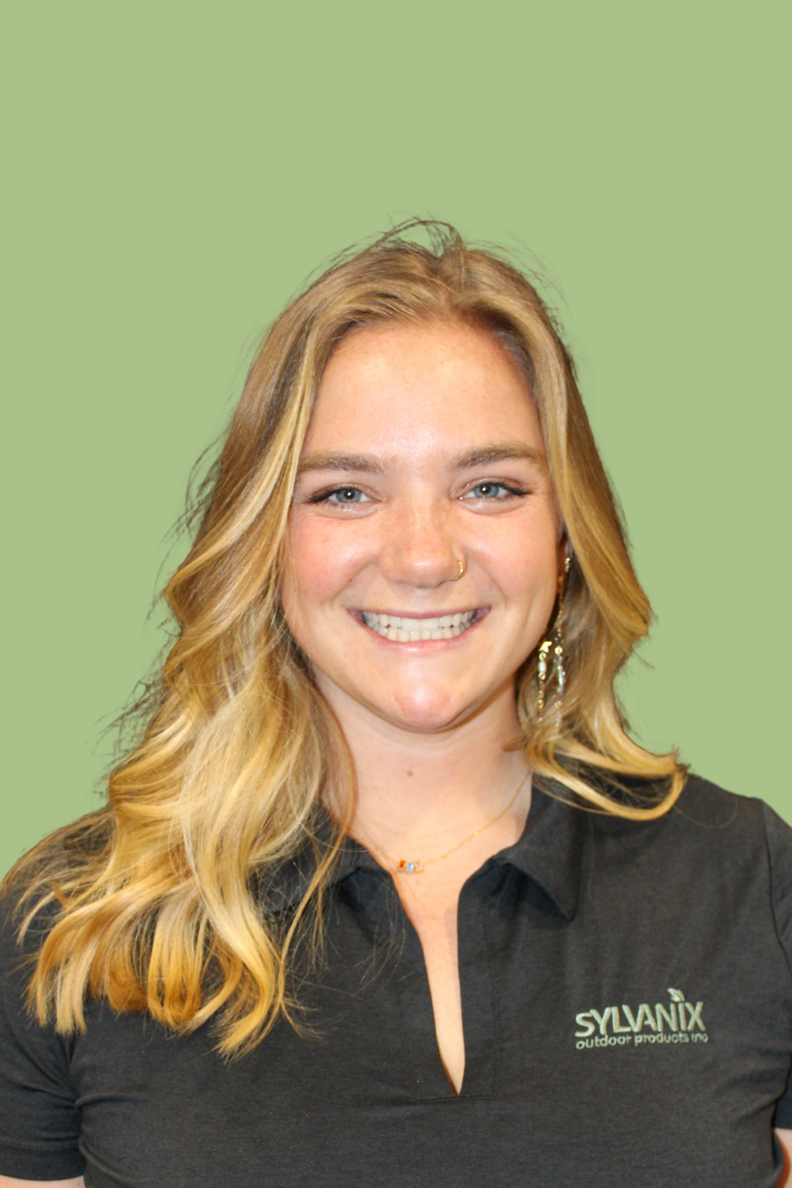 A young woman with blonde wavy hair smiling, wearing a black shirt with 'SYLVANIX outdoor products inc.' logo, earrings, a nose ring, and a necklace, against a plain green background.